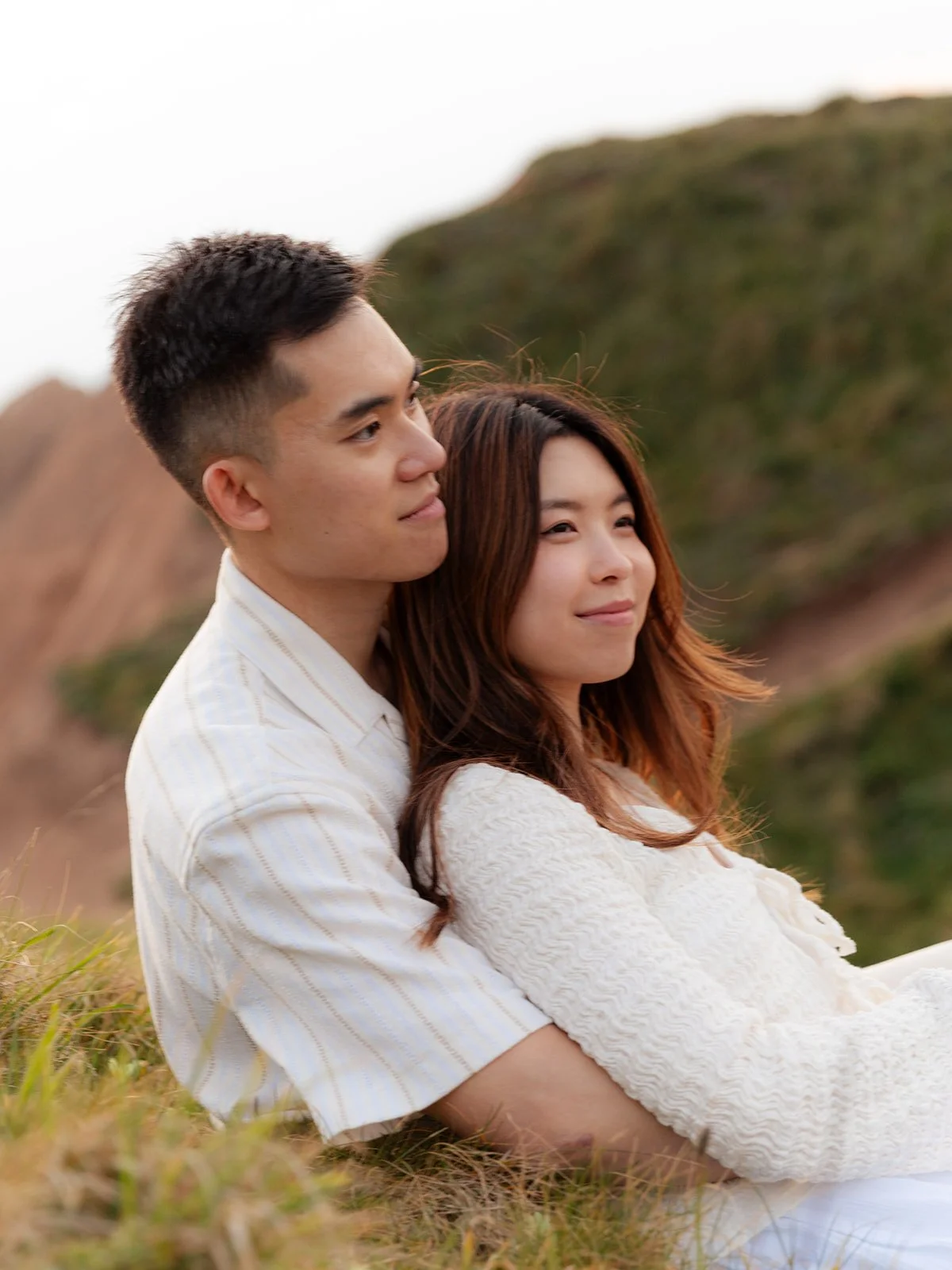 A young couple sitting outdoors on grassy terrain with green hills in the background. The man is embracing the woman from behind as they both look into the distance, appearing content.