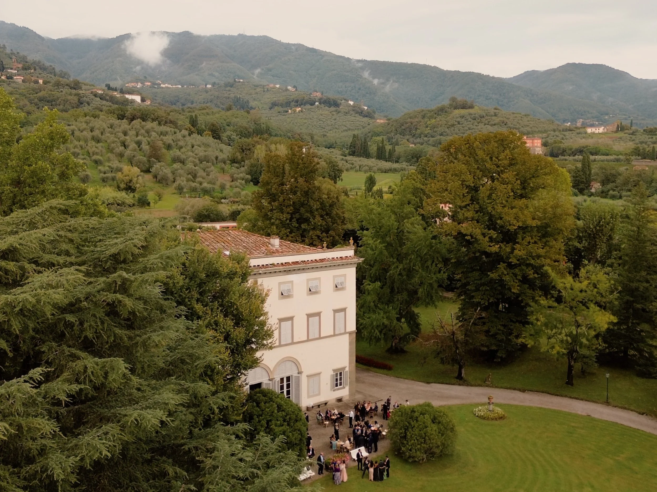 A large group of people gathered outside a white building with arches, trees, and a manicured lawn, with mountains and cloudy sky in the background.