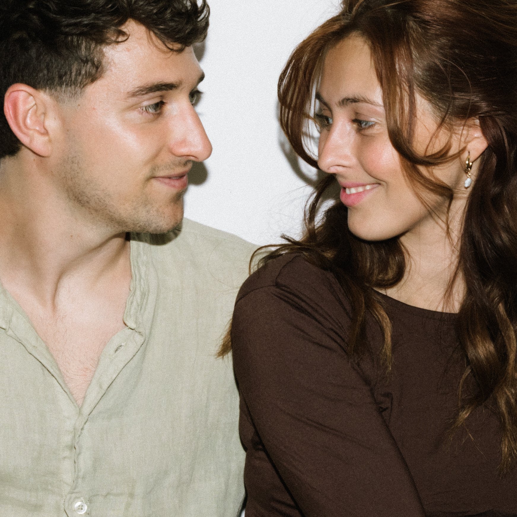 A young man and woman facing each other closely, smiling at each other with their eyes. The man has curly brown hair and a light beige shirt, while the woman has long red hair, earrings, and a brown top. They are mid close-up shot with a plain light background.
