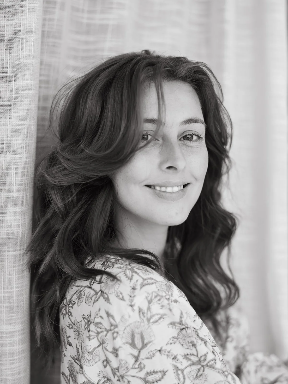 Black and white portrait of a woman with long, wavy hair, smiling, standing near a window with curtains.