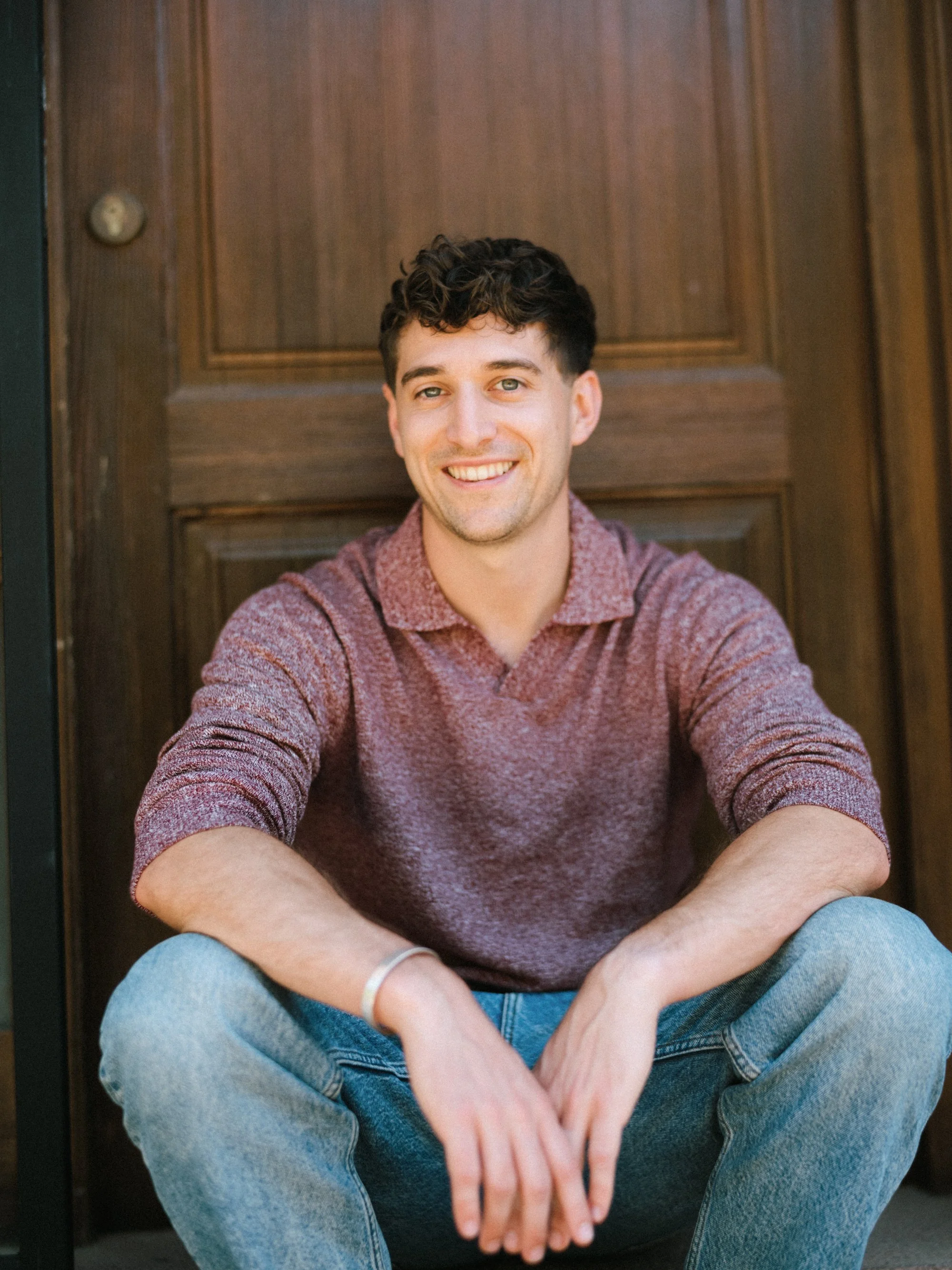 A young man with curly brown hair and light skin smiling, sitting with his legs crossed in front of a wooden door, wearing a maroon long-sleeved shirt and blue jeans.