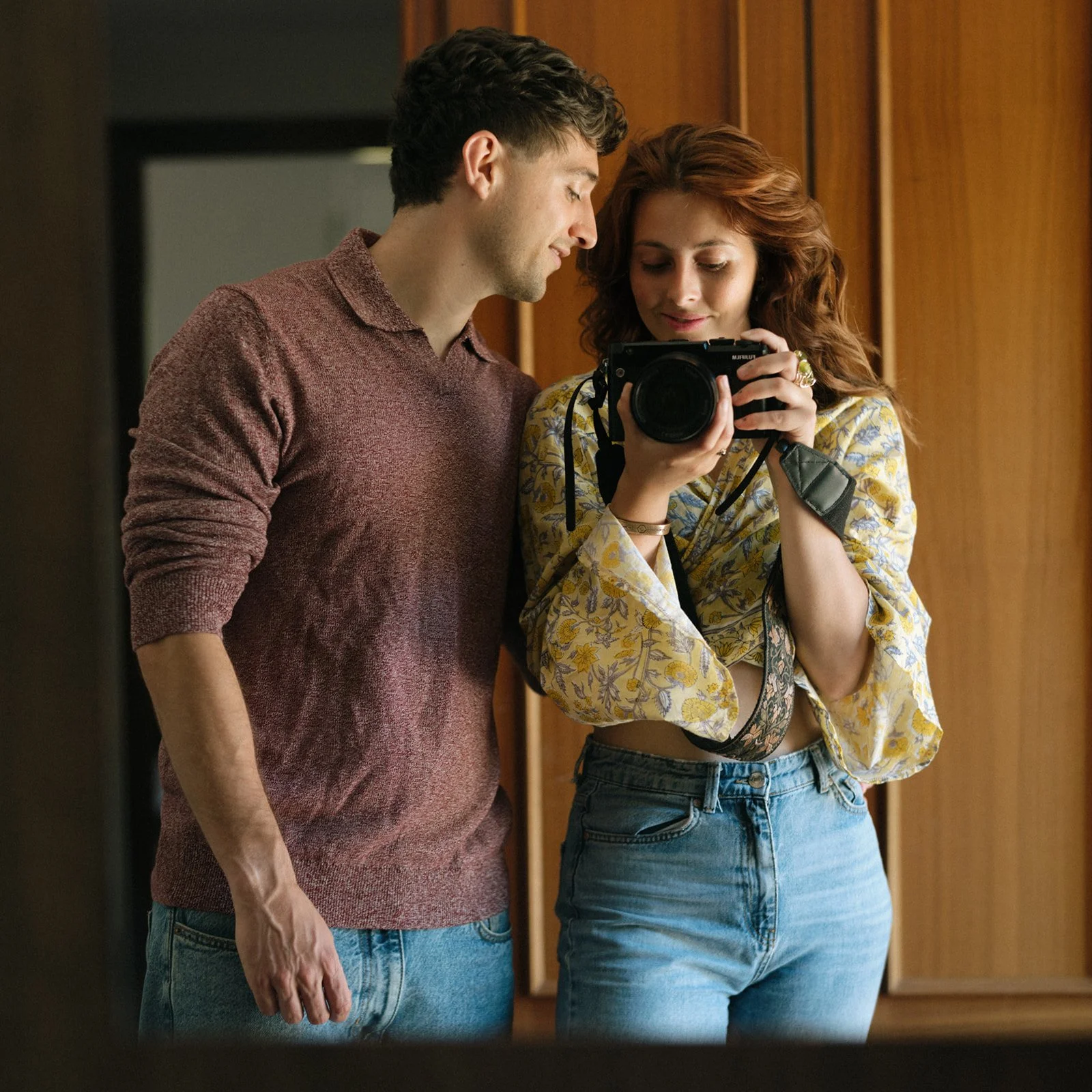 A young man and woman looking at a camera together indoors, with warm wooden paneling in the background.