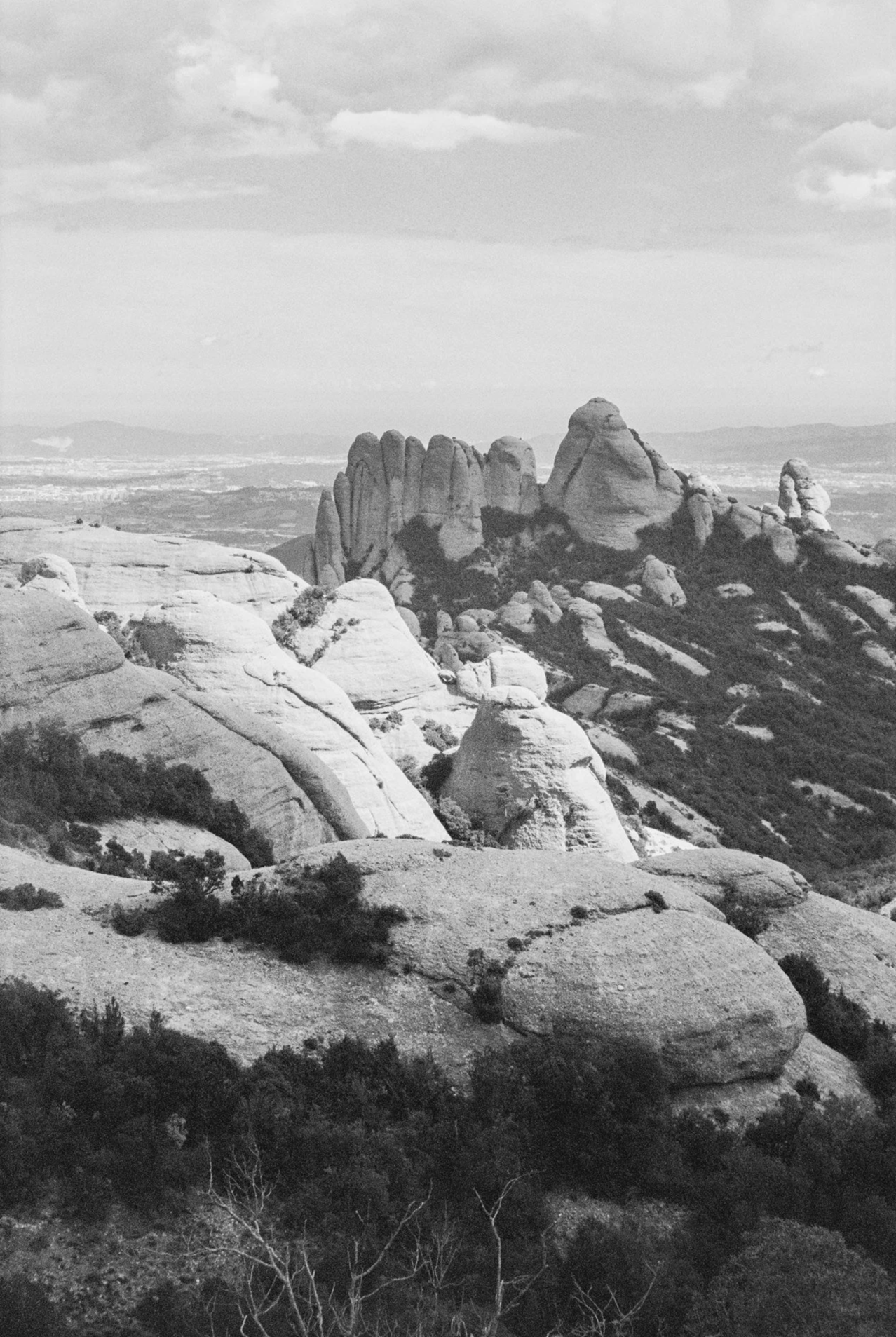 Black and white photo of rocky mountain landscape with large rock formations, sparse vegetation, and a distant view of the horizon with clouds in the sky.