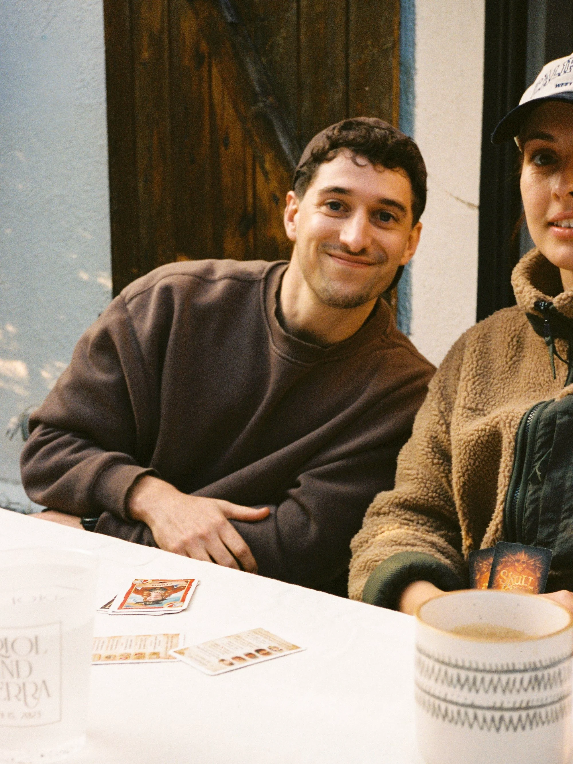 A person with short dark hair wearing a brown hoodie, sitting at a table with playing cards and game pieces. Part of another person wearing a baseball cap and a brown fleece jacket is visible on the right. Items on the table include a deck of cards, a cup, and a stack of poker chips.