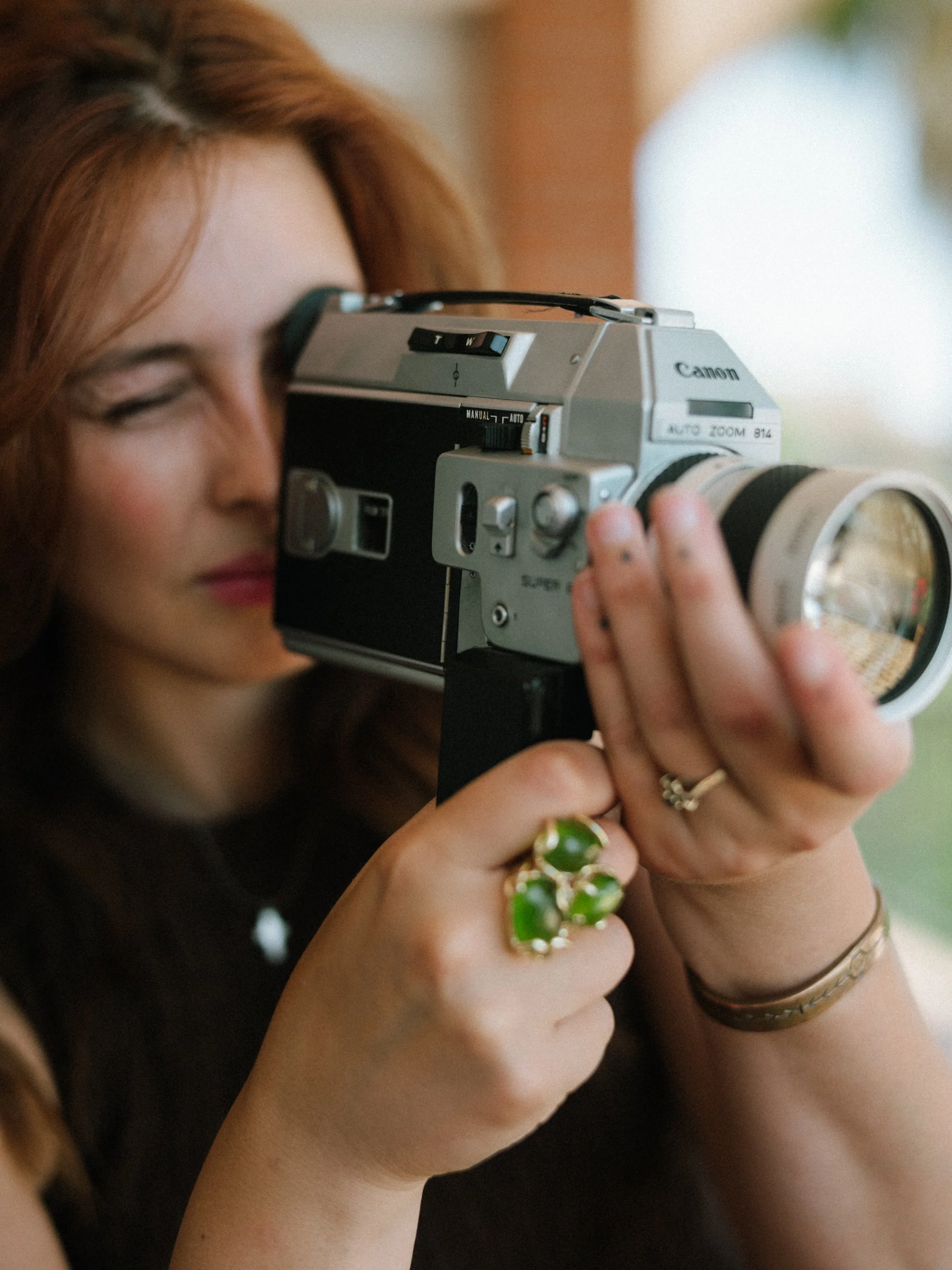 A woman with red hair holding a vintage Canon camera to her face, with her eyes closed, wearing multiple rings and bracelets.