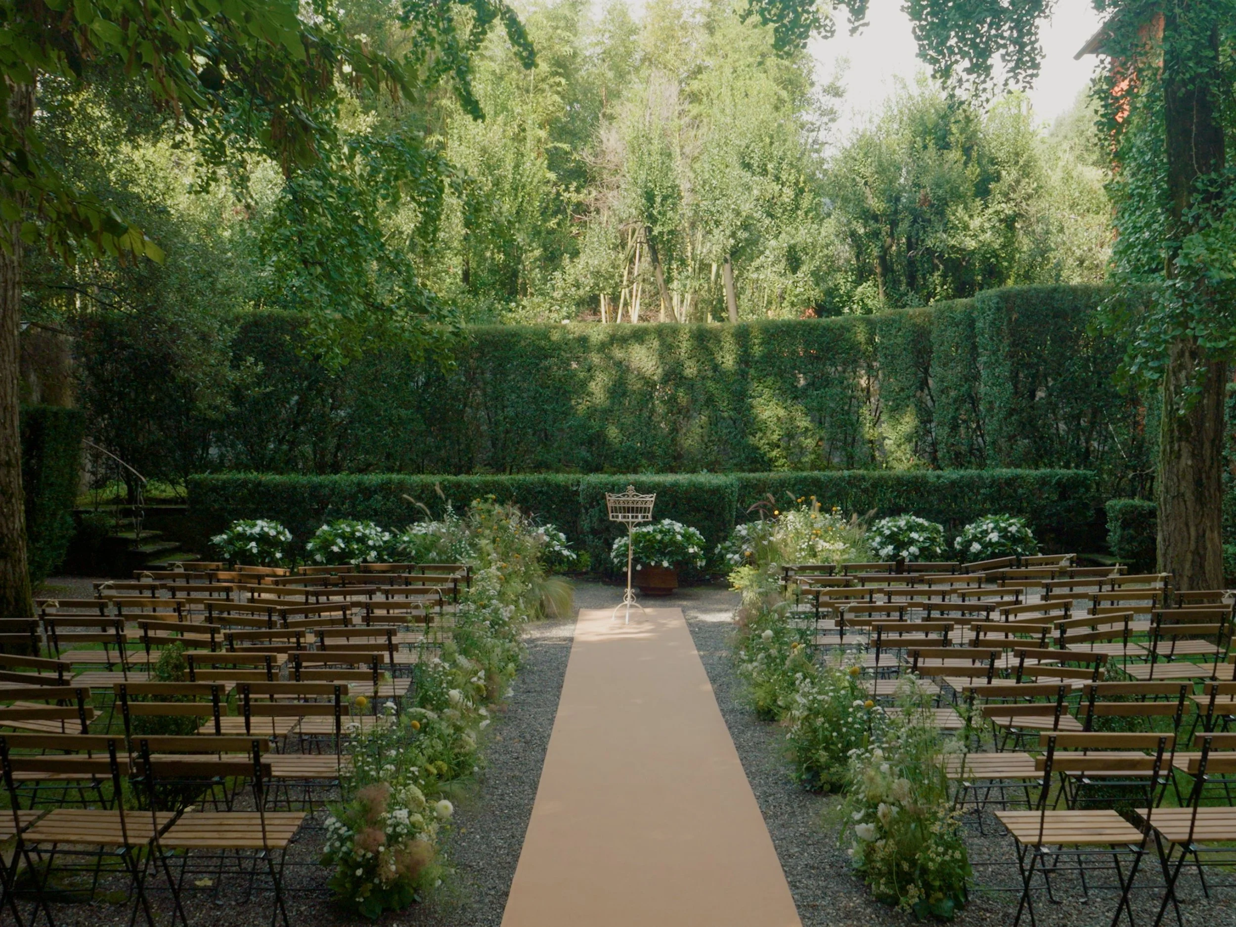 Outdoor wedding ceremony setup with chairs arranged in rows along a central aisle, decorated with flowers, in a lush green garden with tall trees and hedges.