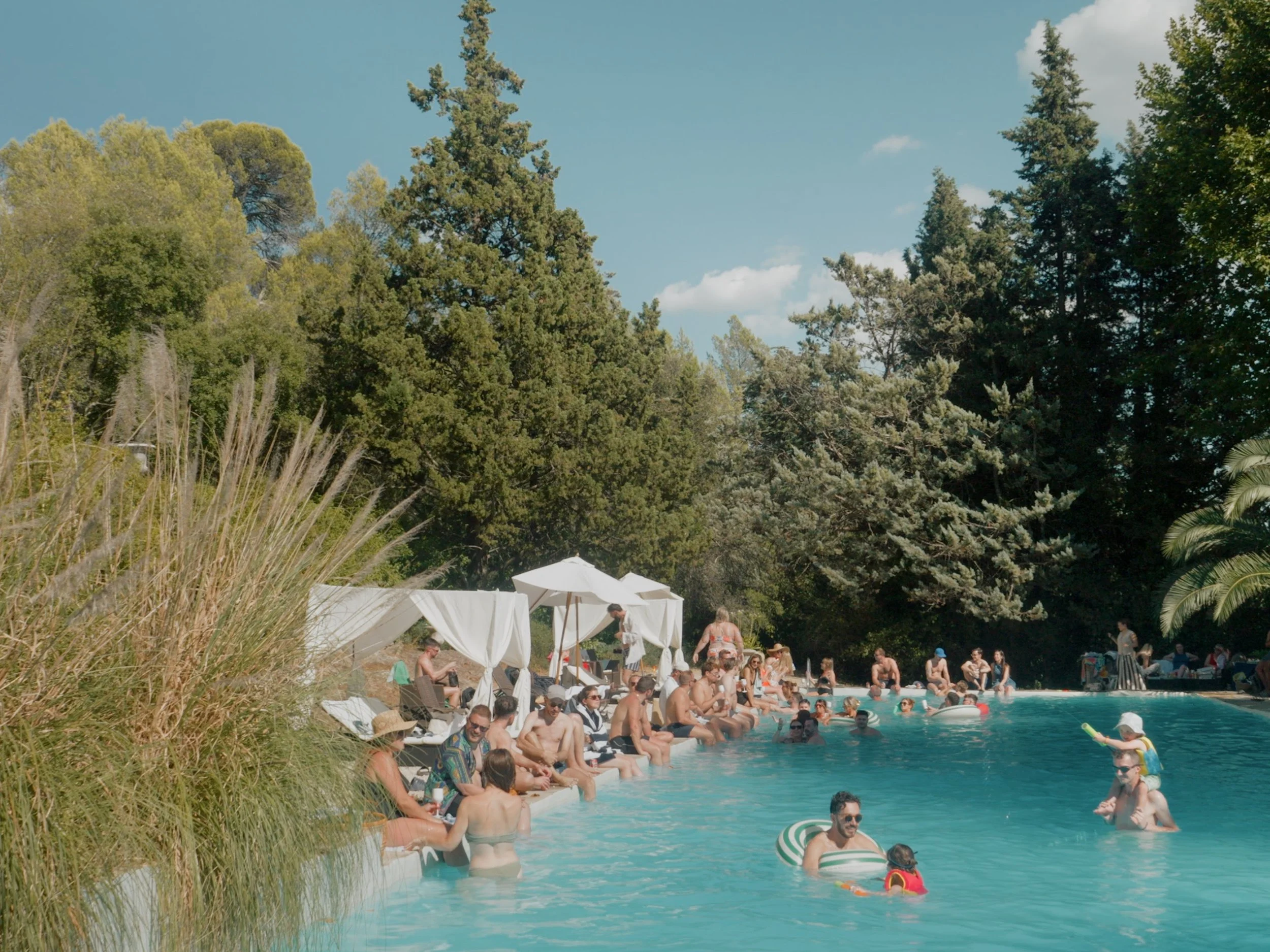 People swimming and lounging by a pool surrounded by green trees and umbrellas on a sunny day.