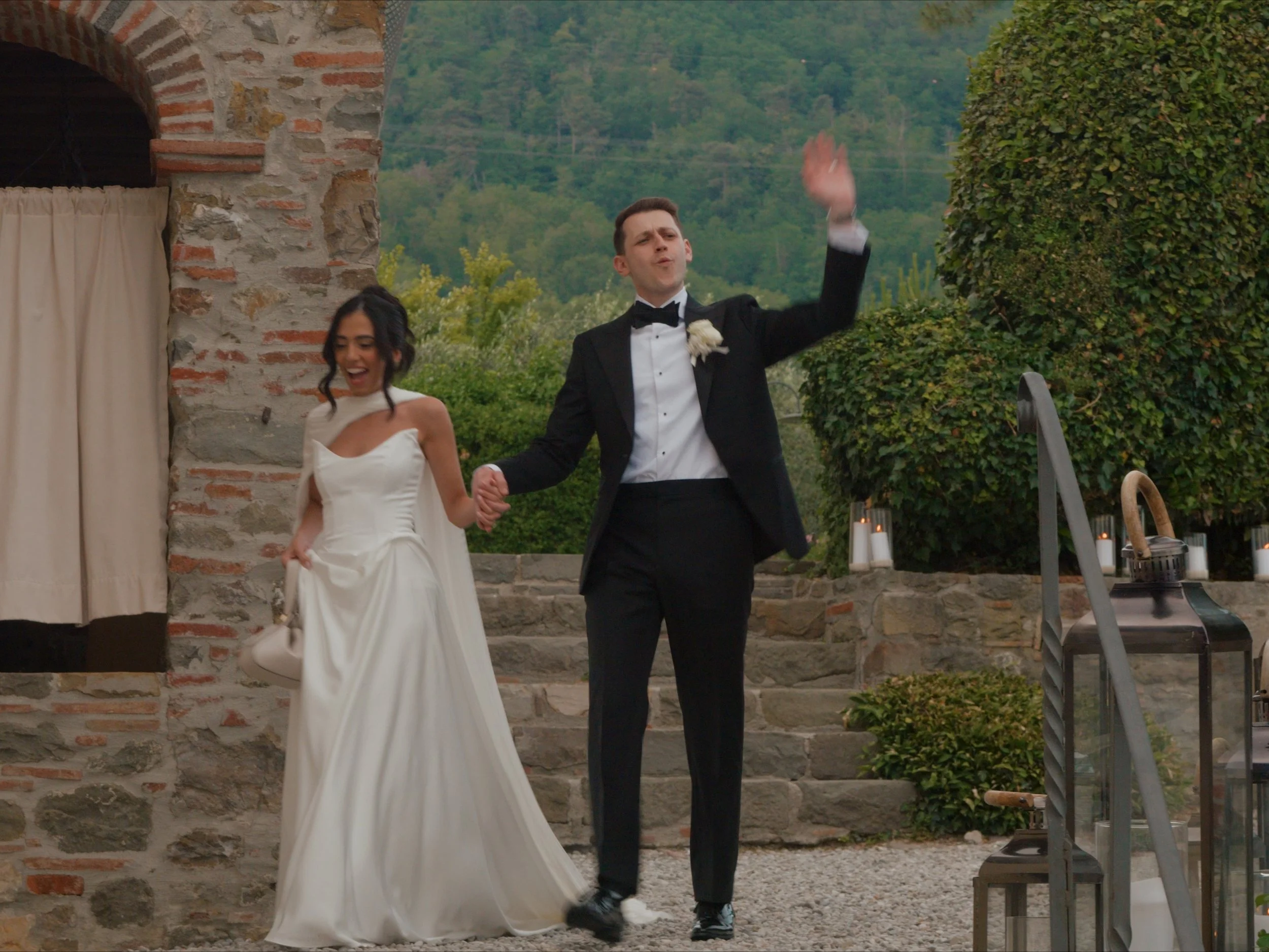 A newlywed couple holding hands and dancing outdoors. The groom is wearing a tuxedo with a bowtie and boutonniere, and the bride is wearing a white wedding dress with a strapless neckline. They are smiling and appear to be enjoying their moment. There is a stone building or wall and lush green trees in the background.