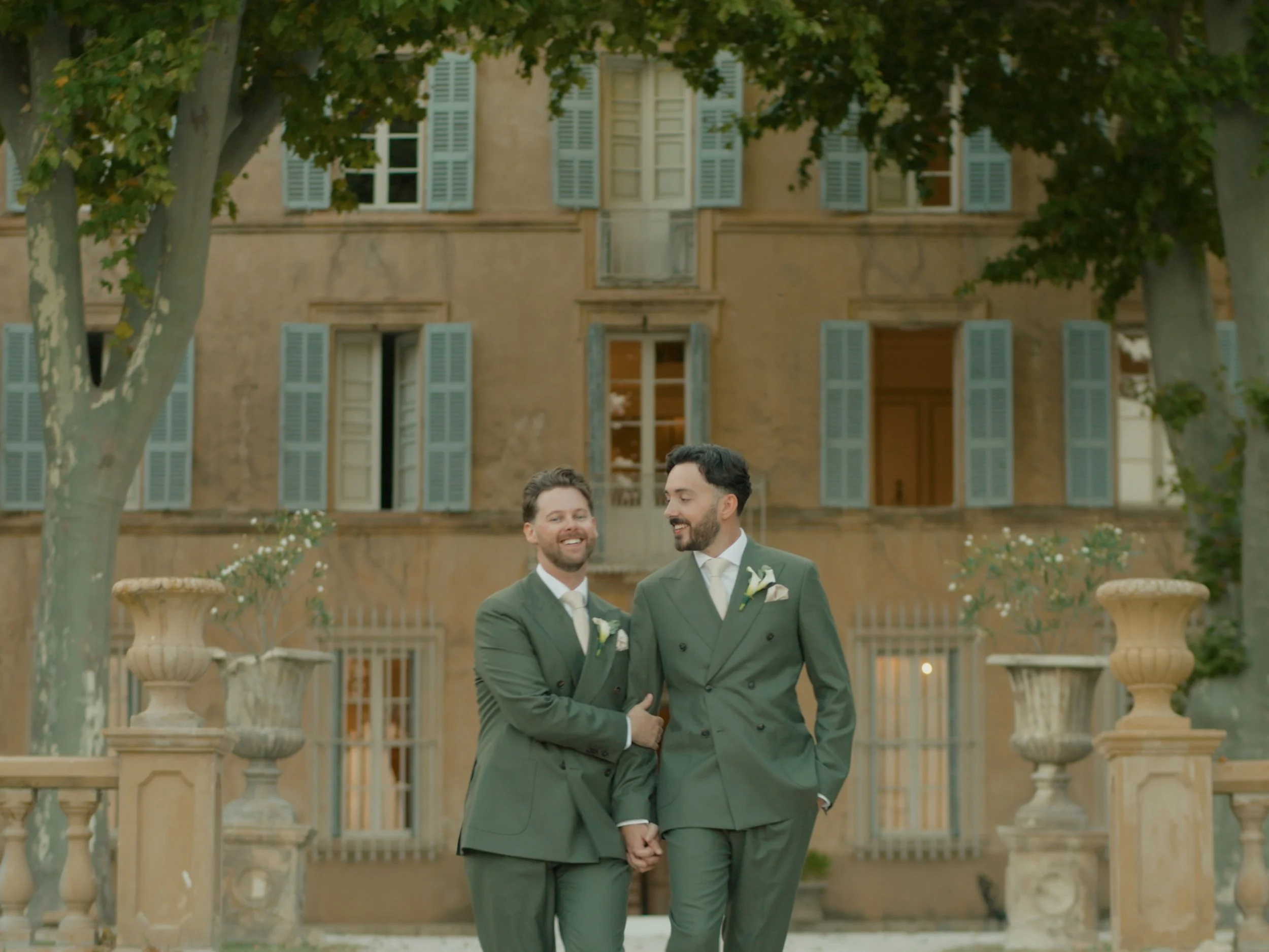 Two men in formal suits holding hands and smiling outdoors in front of Chateau Robernier with multiple windows, surrounded by trees and stone planters.