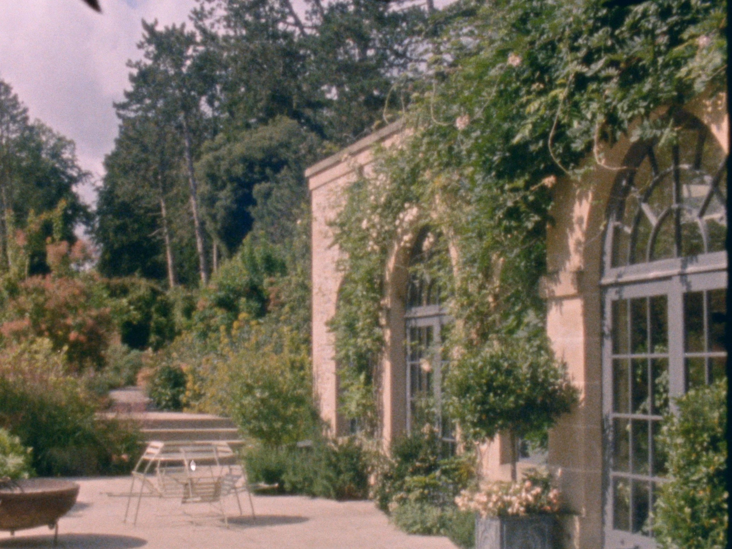 A view of Middleton Lodge Estate wedding venue with multiple windows, surrounded by trees with green foliage. In the foreground, there is a patio area with a glass table and chairs.