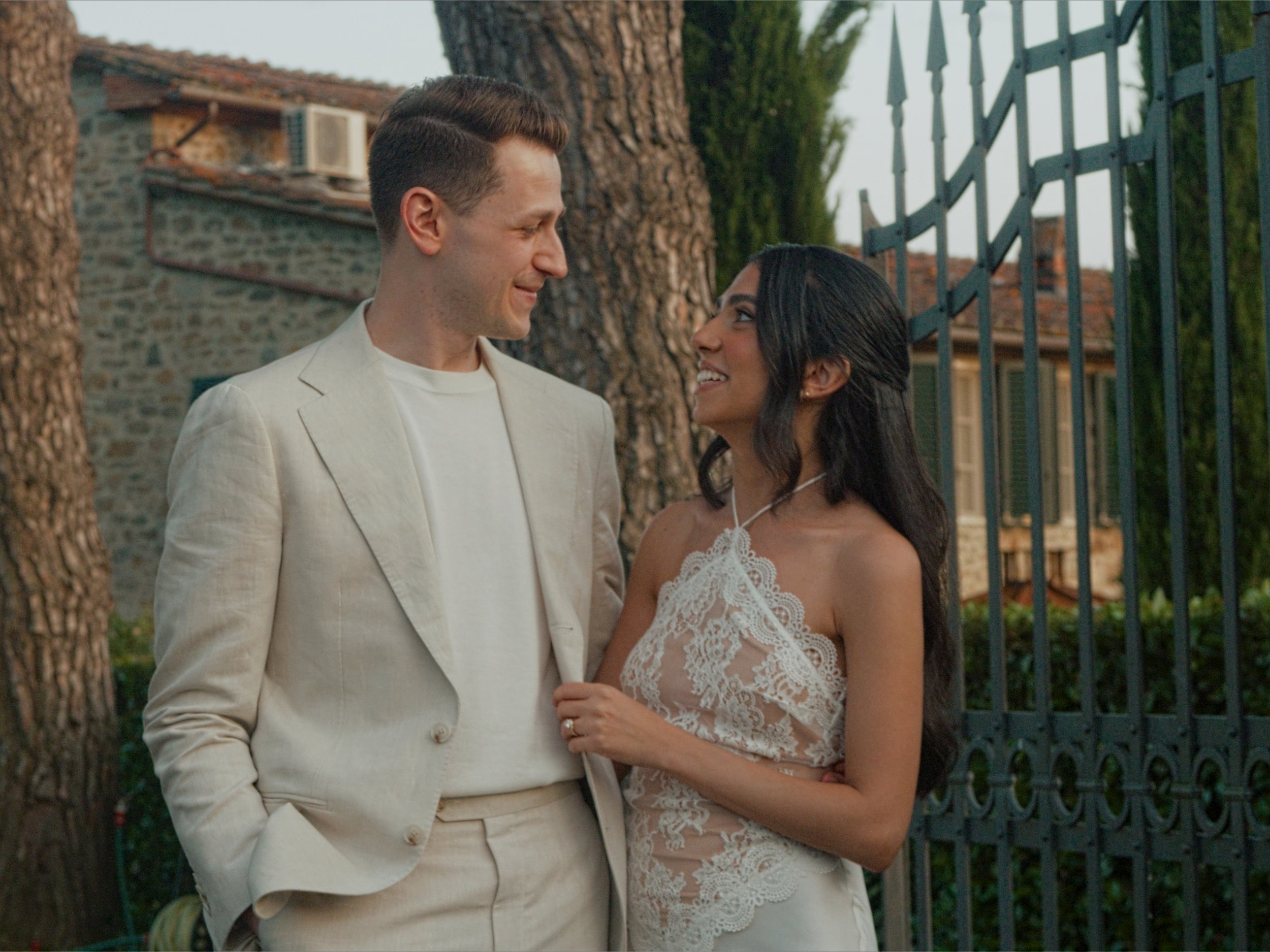A man and woman standing close and smiling at each other outdoors near trees and a metal fence, dressed in wedding attire.