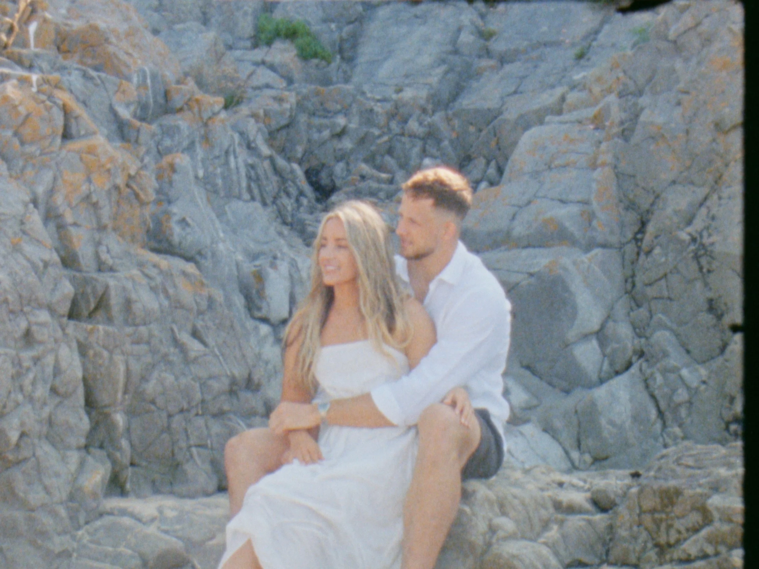 A couple sitting together on the rocks at a beach, smiling and enjoying the outdoor scene in a couple portrait video session