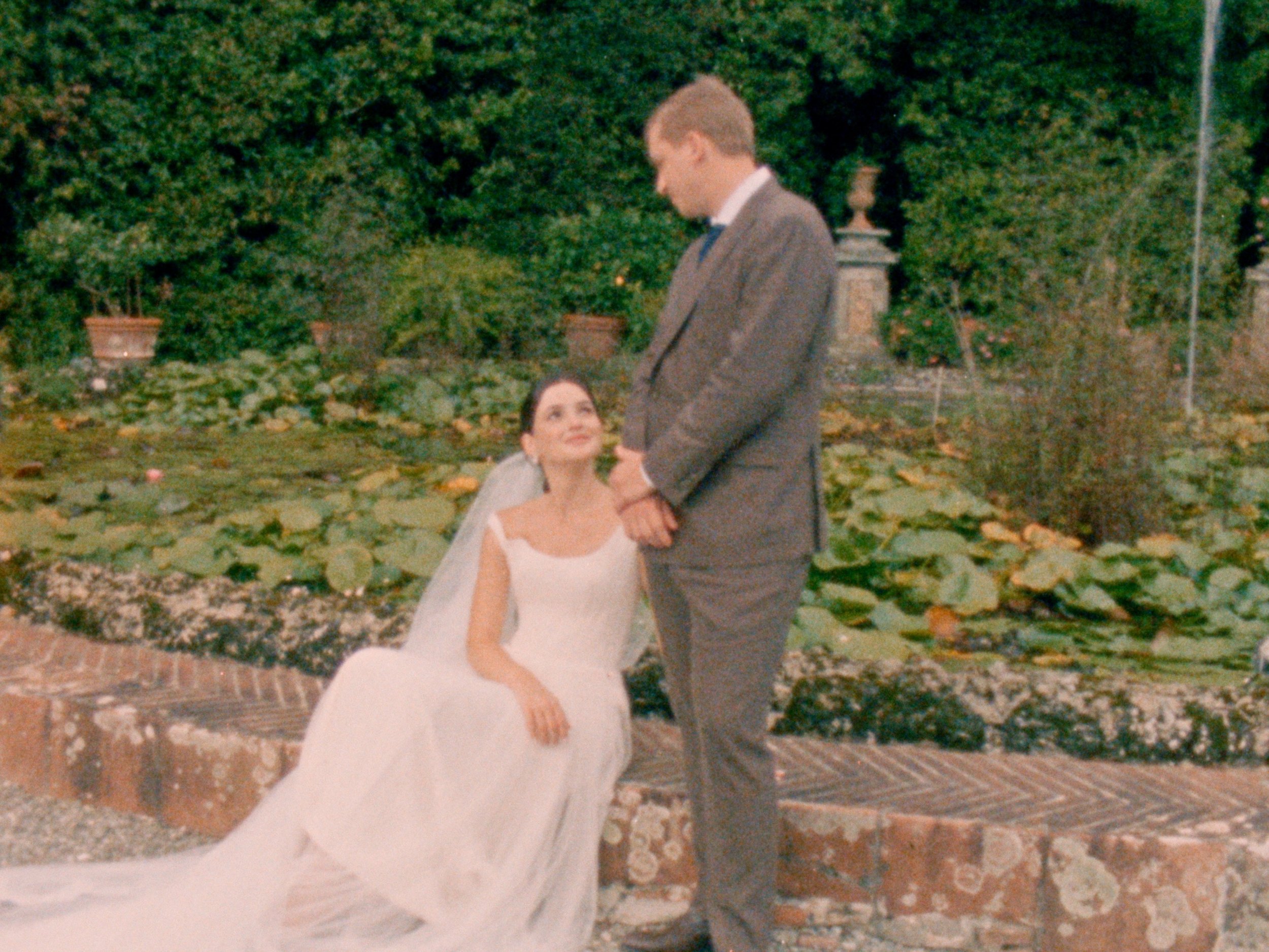 A woman in a white wedding dress sitting on a low wall, holding the hand of a man in a gray suit, with a garden and pond with lily pads in the background.