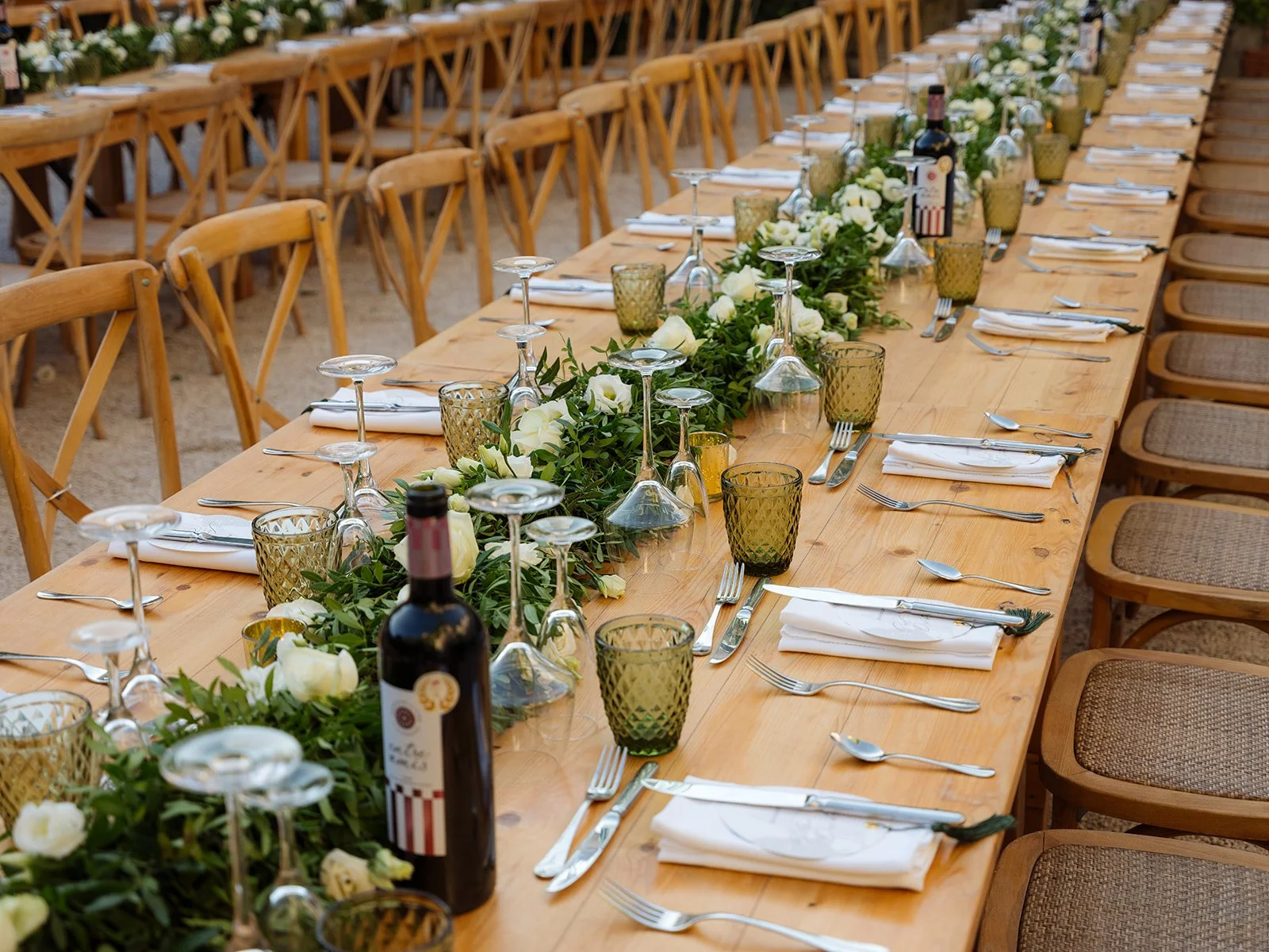 Long rectangular wooden wedding dining table set for a formal wedding at Chateau Robernier event with white napkins, silverware, green glass cups, wine glasses upside down, and a floral centerpiece with white roses and greenery.