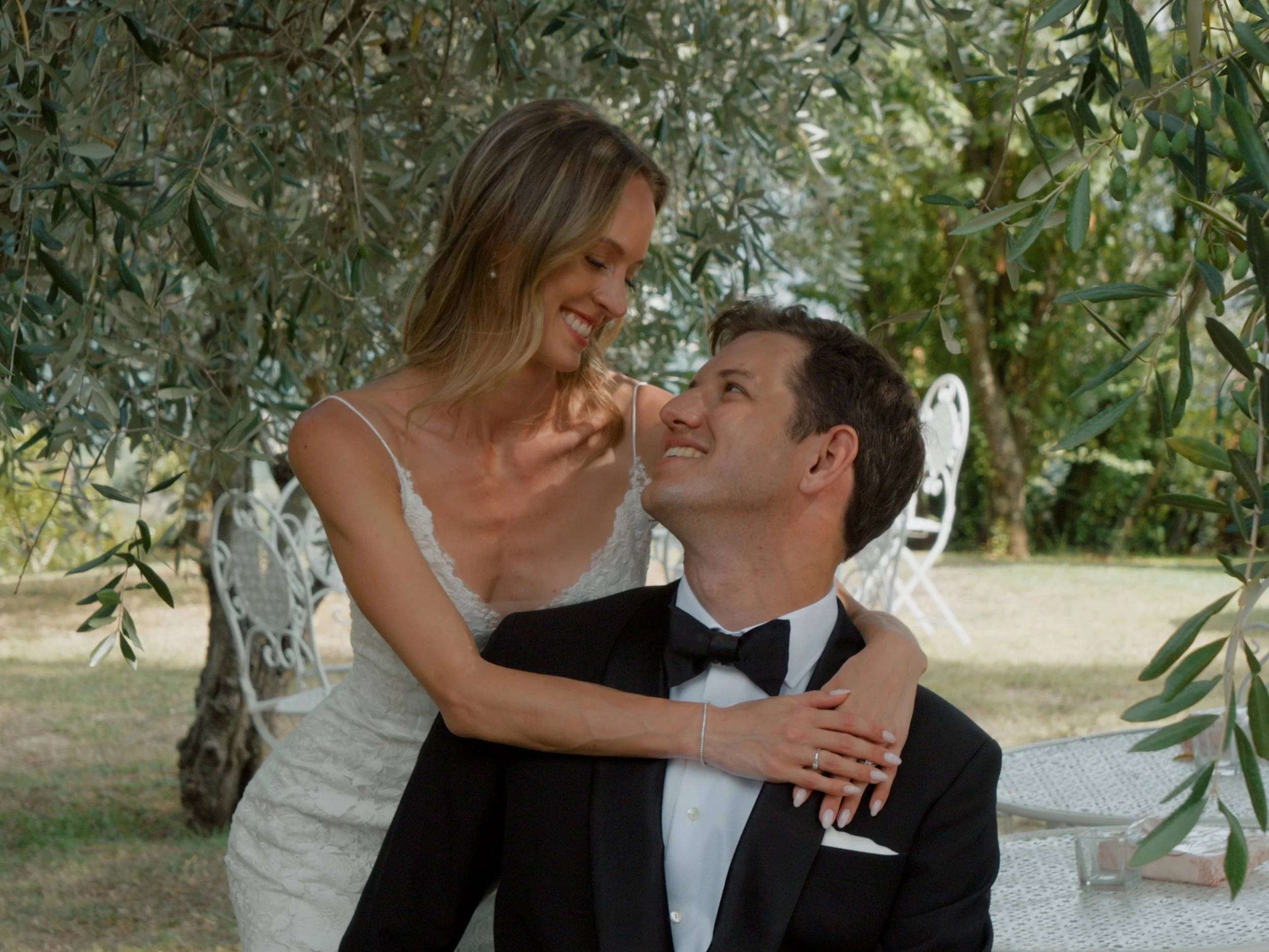 A happy couple seated outdoors among greenery, with the woman wearing a white dress and the man in a tuxedo, gazing at each other affectionately.