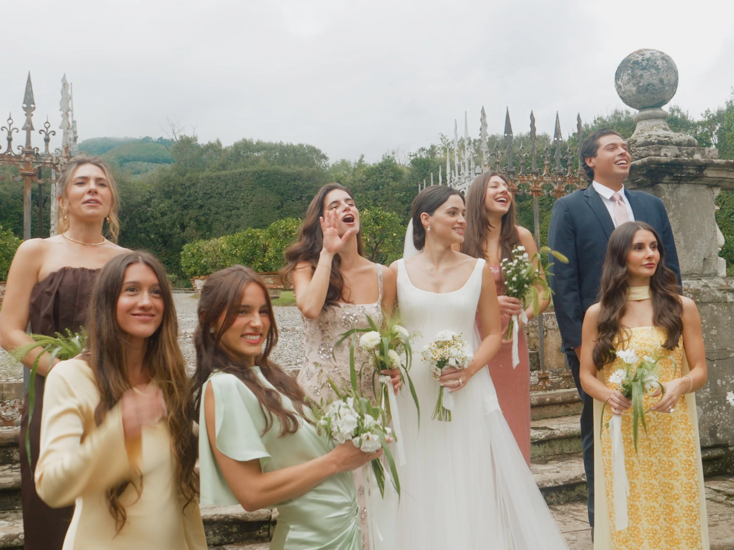 Group of women and one man in formal attire smiling and holding bouquets, standing outdoors near stone steps and ornate iron fence, with trees and cloudy sky in background.