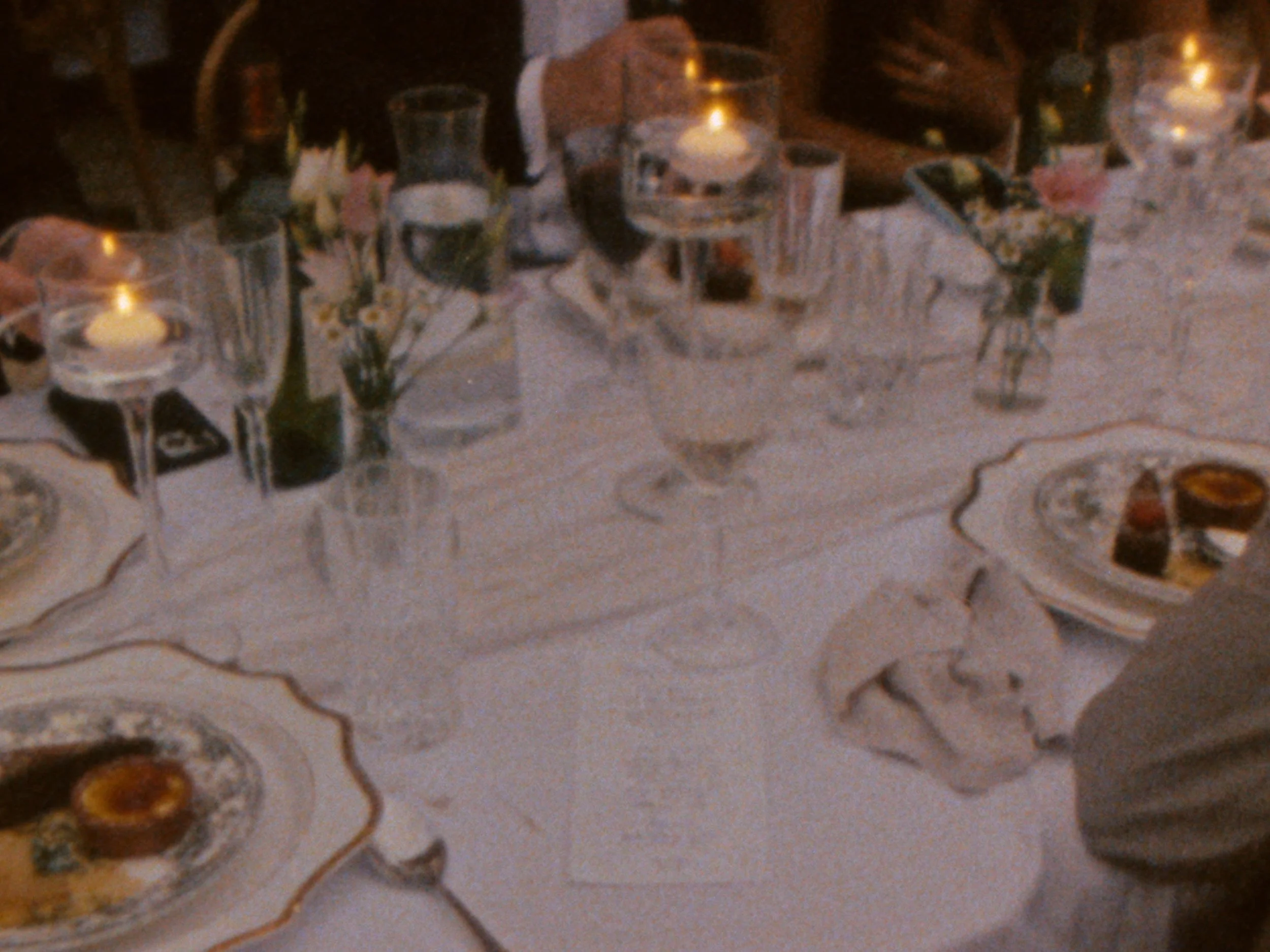 A close-up view of a decorative, possibly vintage, table setting with glasses, floral arrangements, and dishes on a tablecloth, with warm candle lighting at a wedding evening reception in Provence, France