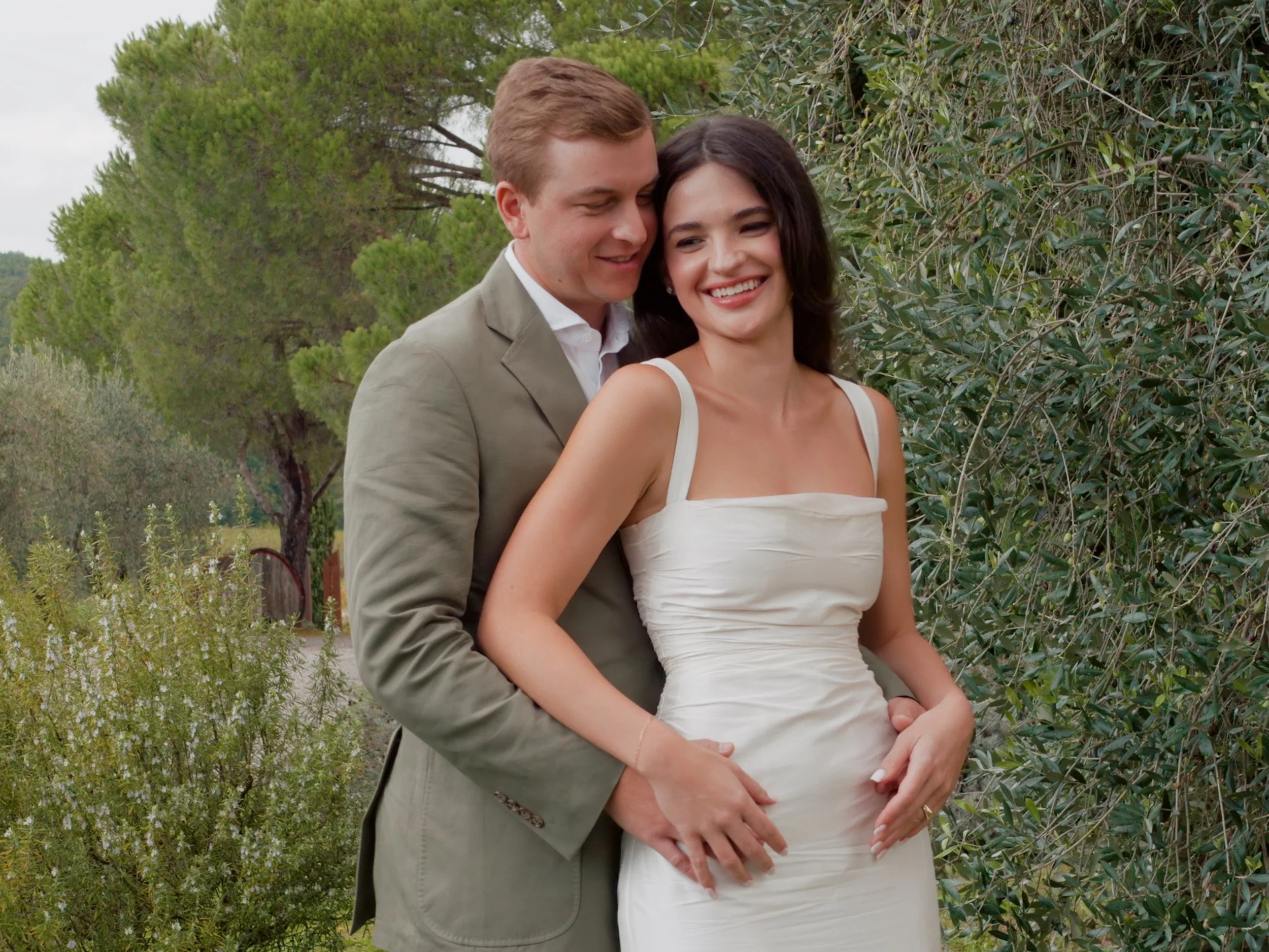 A couple smiling and embracing outdoors among trees and greenery, with the woman in a white dress and the man in a light gray suit. Taken at Villa Grabau, Lucca