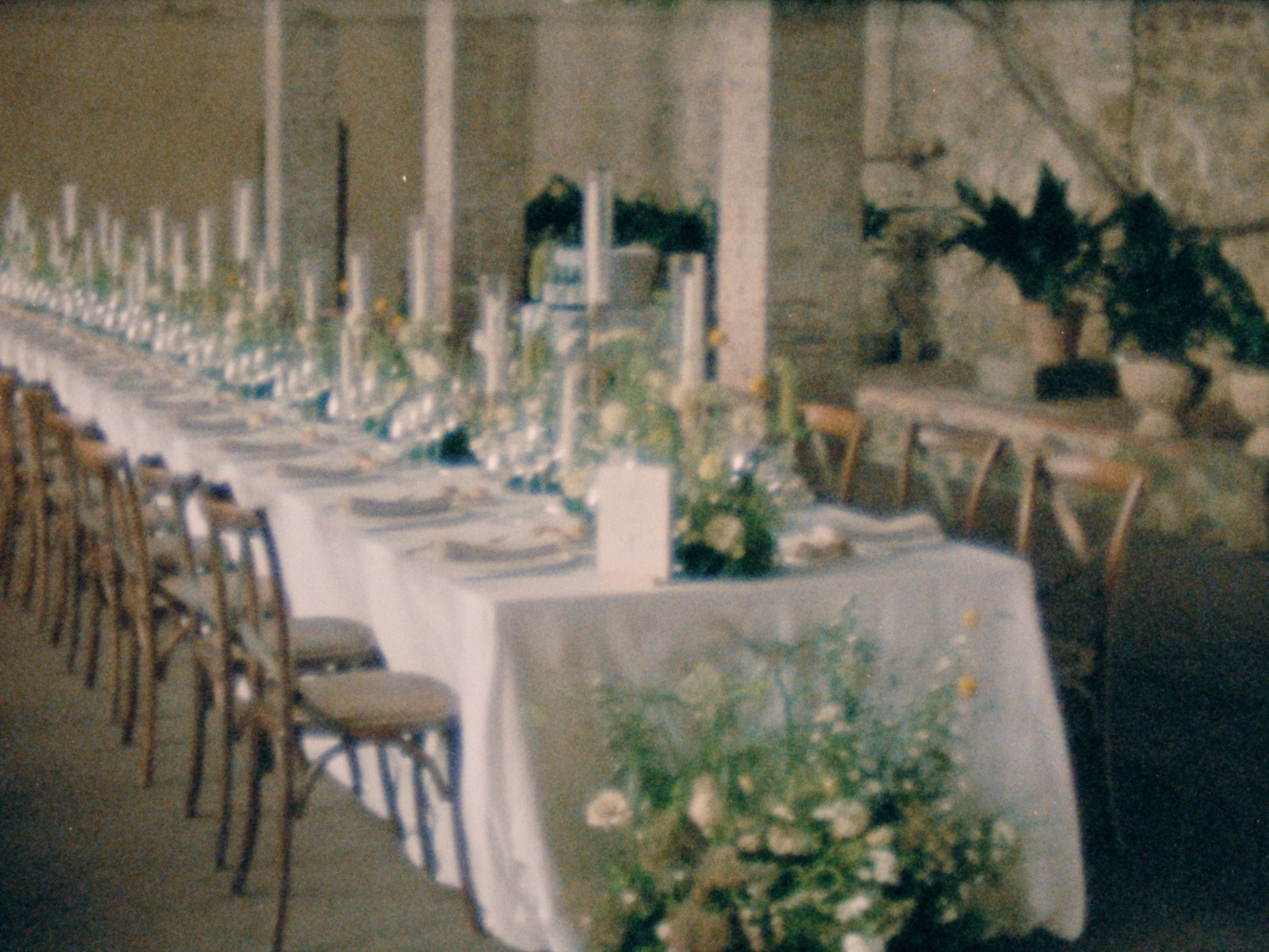 A long wedding dining table with wooden chairs on both sides, set outdoors with greenery and string lights in the background at Villa Grabau