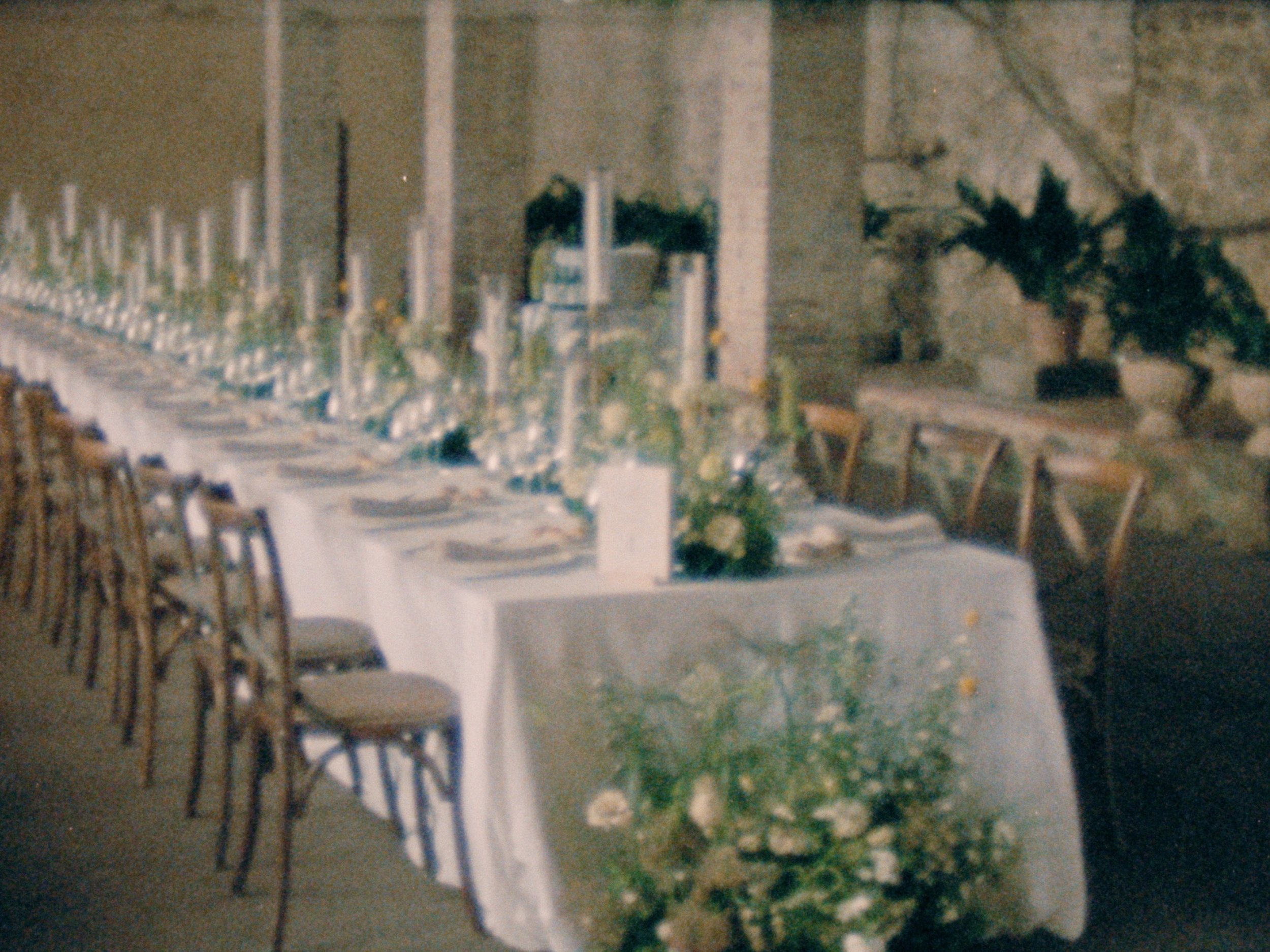 A banquet table outdoors with chairs, tablecloth, and menu cards, surrounded by greenery and a building in the background.