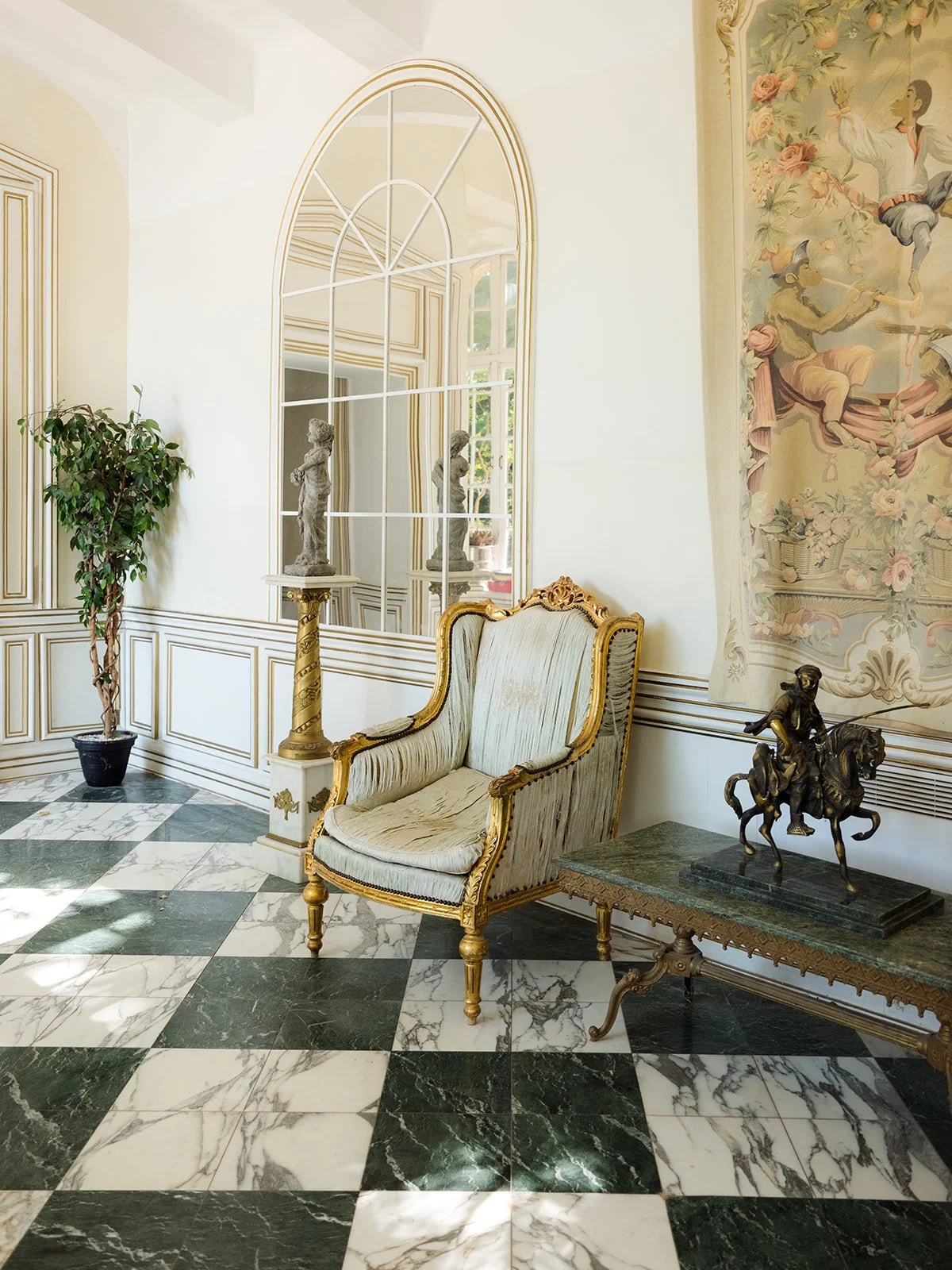 Elegant room with black and white marble checkered floor, ornate gold-trimmed armchair, potted plant, tall mirror, sculptures, and a large tapestry on the wall.