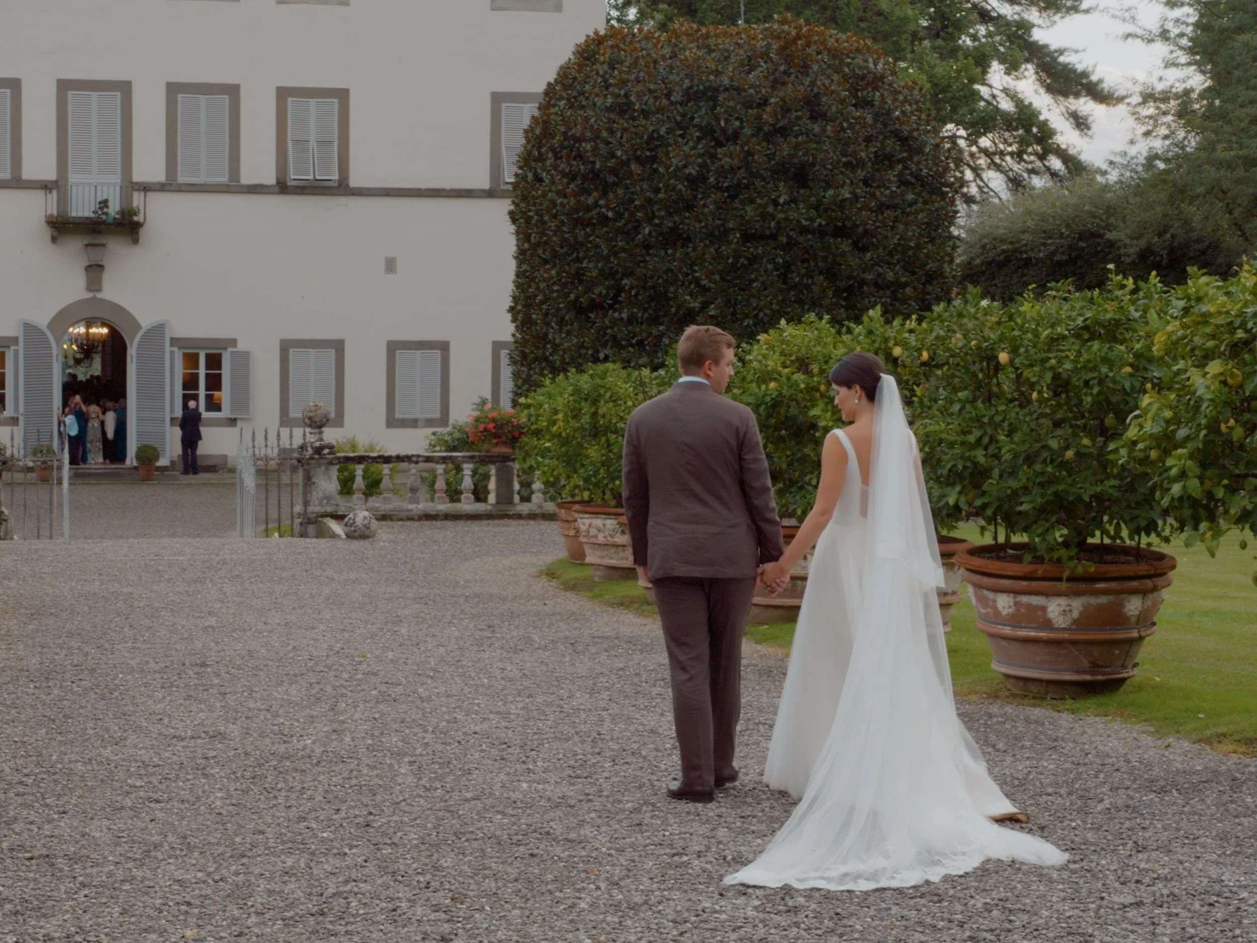 A bride and groom holding hands outdoors in front of a large white house with tall windows and shutters, surrounded by potted plants and greenery.