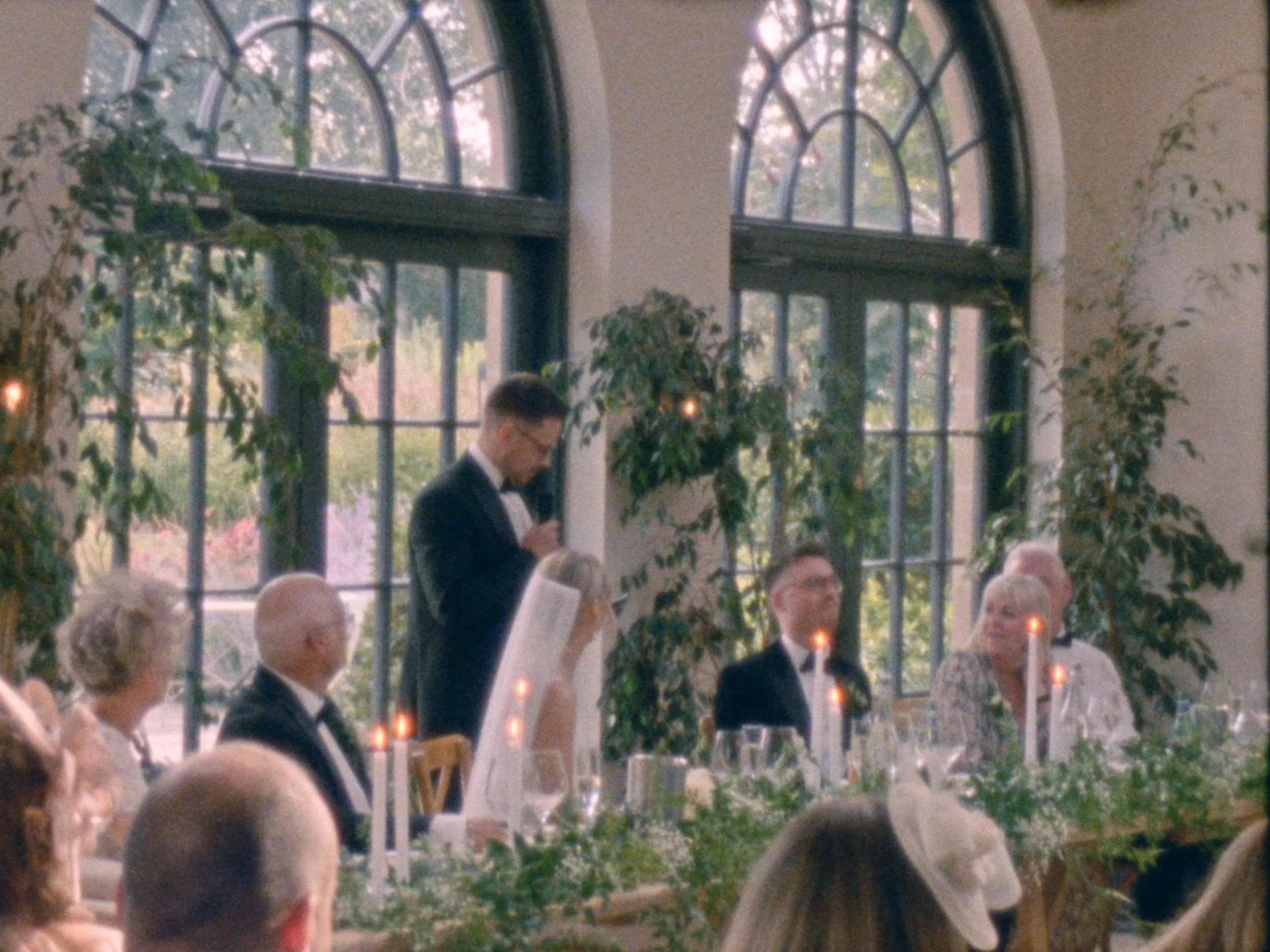 People attending a formal event or reception inside a room with large arched windows and greenery outside. The scene shows a man in a tuxedo standing and looking down while other people are seated, with some candles on the table.