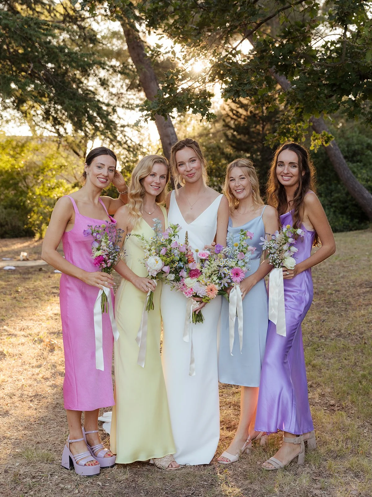 A group of five women in pastel-colored dresses holding bouquets of flowers, standing outdoors in a park during sunset.