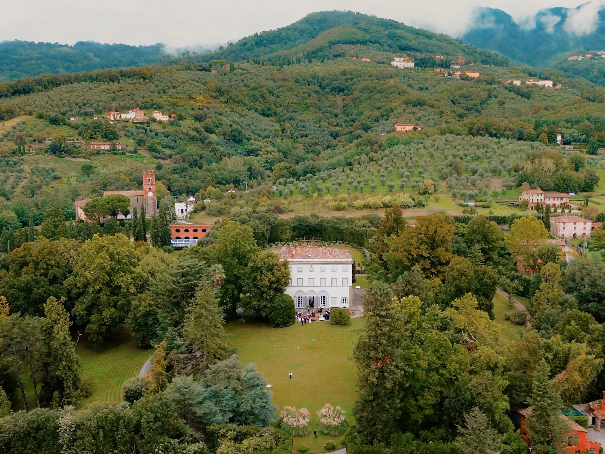 A scenic hillside landscape with Villa Grabau, Lucca, dense greenery, and a small building with an outdoor gathering area, set against a backdrop of forested mountains under a cloudy sky.