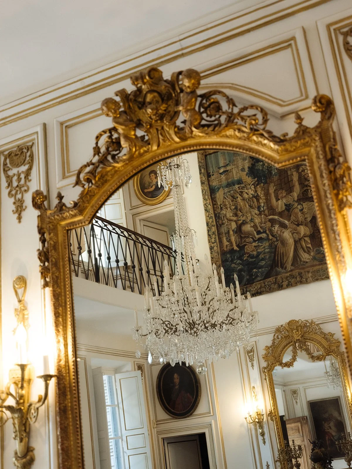An ornate gold-framed mirror reflecting a crystal chandelier, a staircase with intricate railing, classical paintings, a tapestry, and gilded wall decorations in a luxurious room inside Chateau Robernier