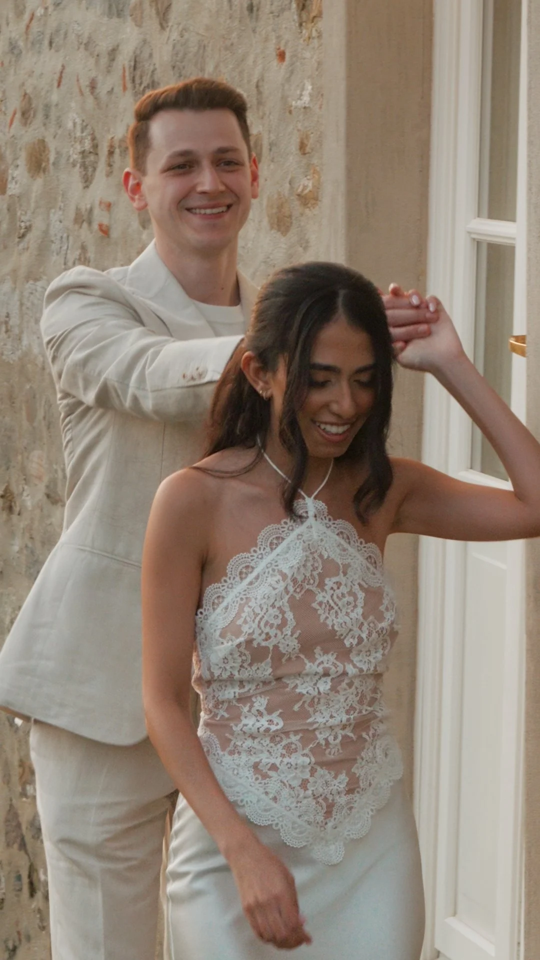 A young man in a beige suit and a young woman in a white lace top are smiling and playfully holding hands above her head in a cozy indoor setting at Casale de Pasquinelli