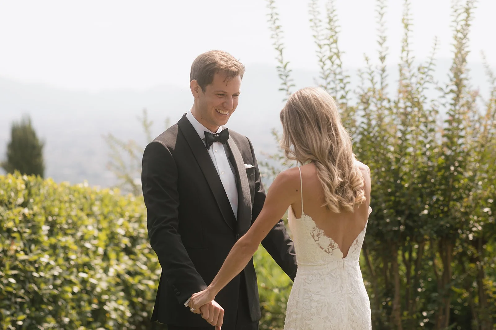 A bride and groom holding hands during their wedding ceremony outdoors, with the groom in a tuxedo and the bride in a white lace gown, surrounded by greenery.