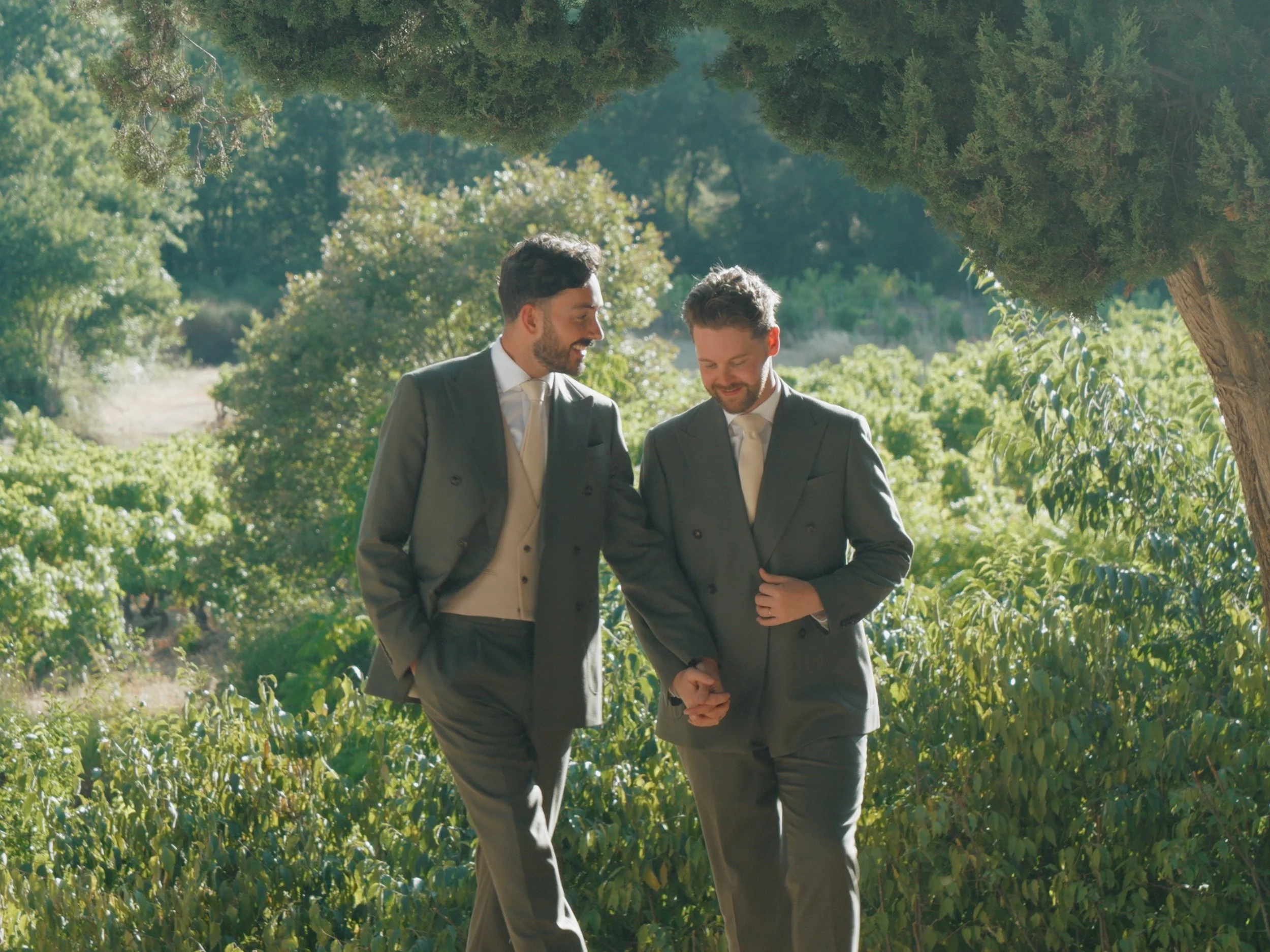 Two men in suits are walking together outdoors in a green, wooded area, smiling and engaging in conversation.
