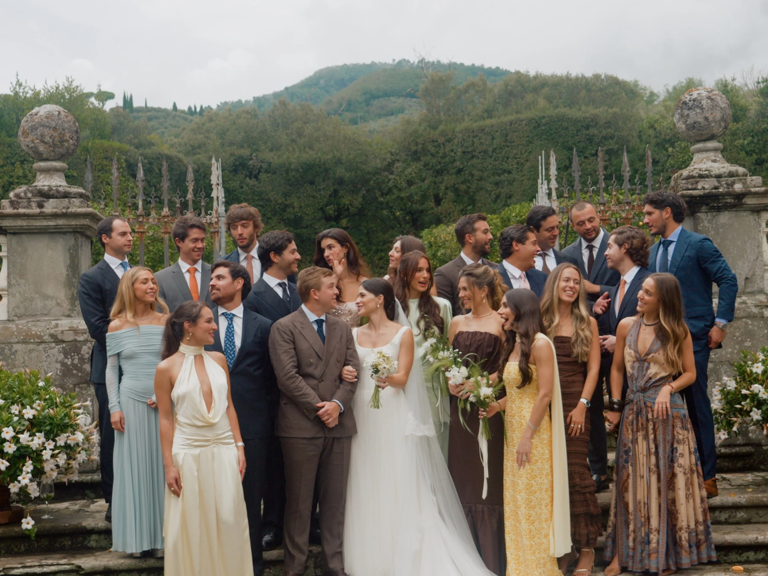 A large group of people dressed in formal wedding attire, gathered outdoors in front of an iron fence and stone columns, with a lush green hill in the background at villa grabau
