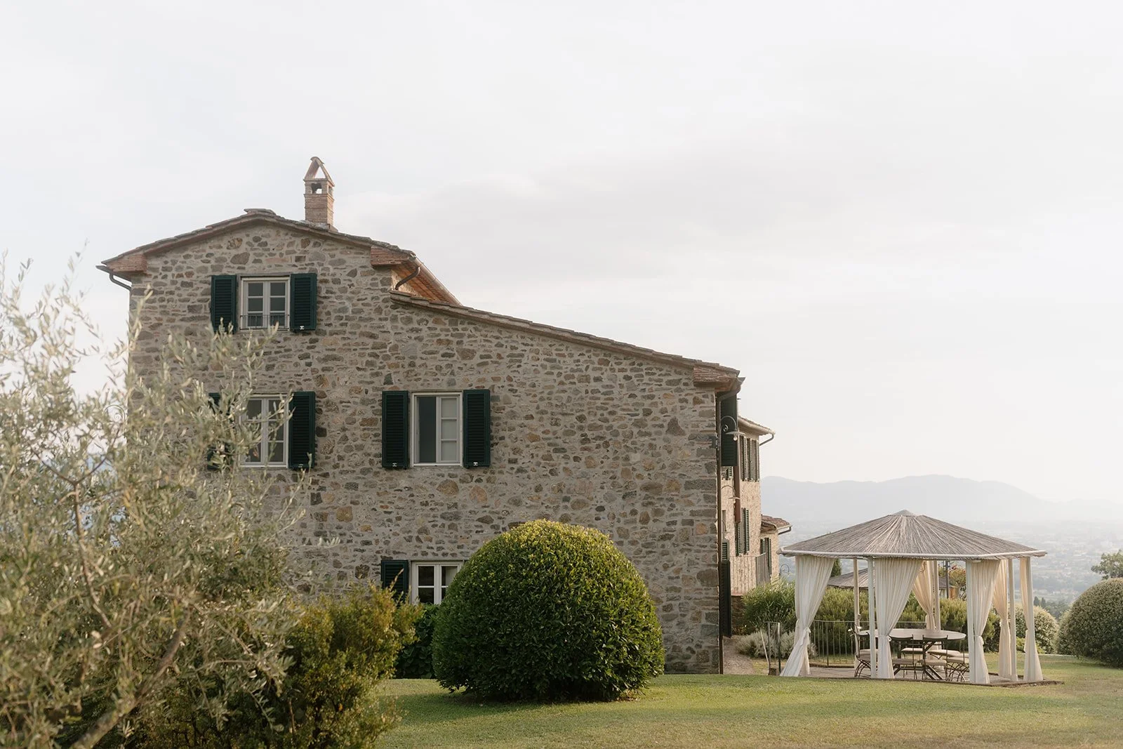 Stone house with green shutters, a gazebo with curtains, and landscaping with bushes and trees.