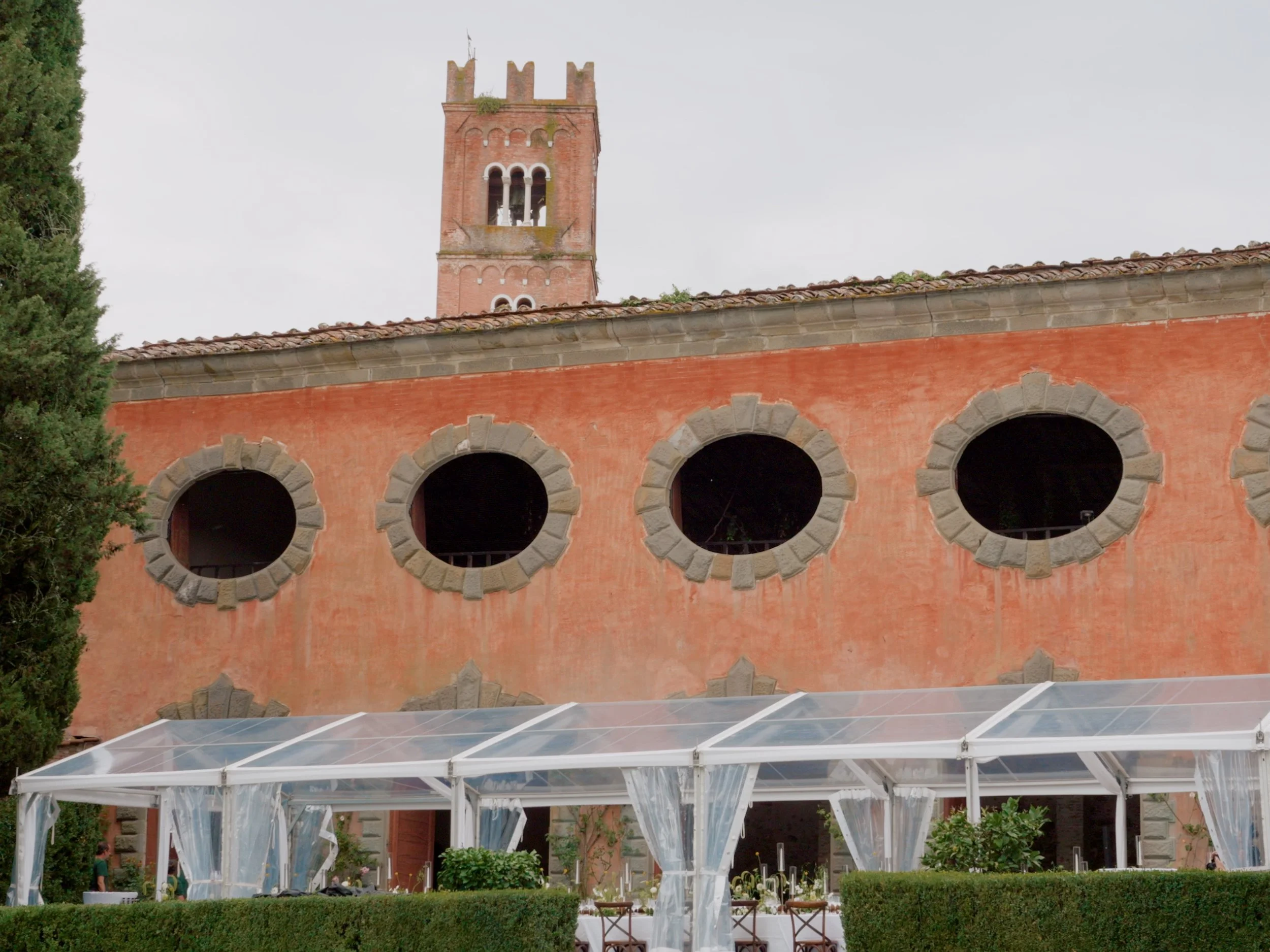 A historic red brick building with a tall clock tower featuring arched windows and crenellations on top. In front, there is a transparent glass structure, possibly a greenhouse or conservatory, with tables and chairs underneath. Greenery surrounds the area, including trees and bushes.