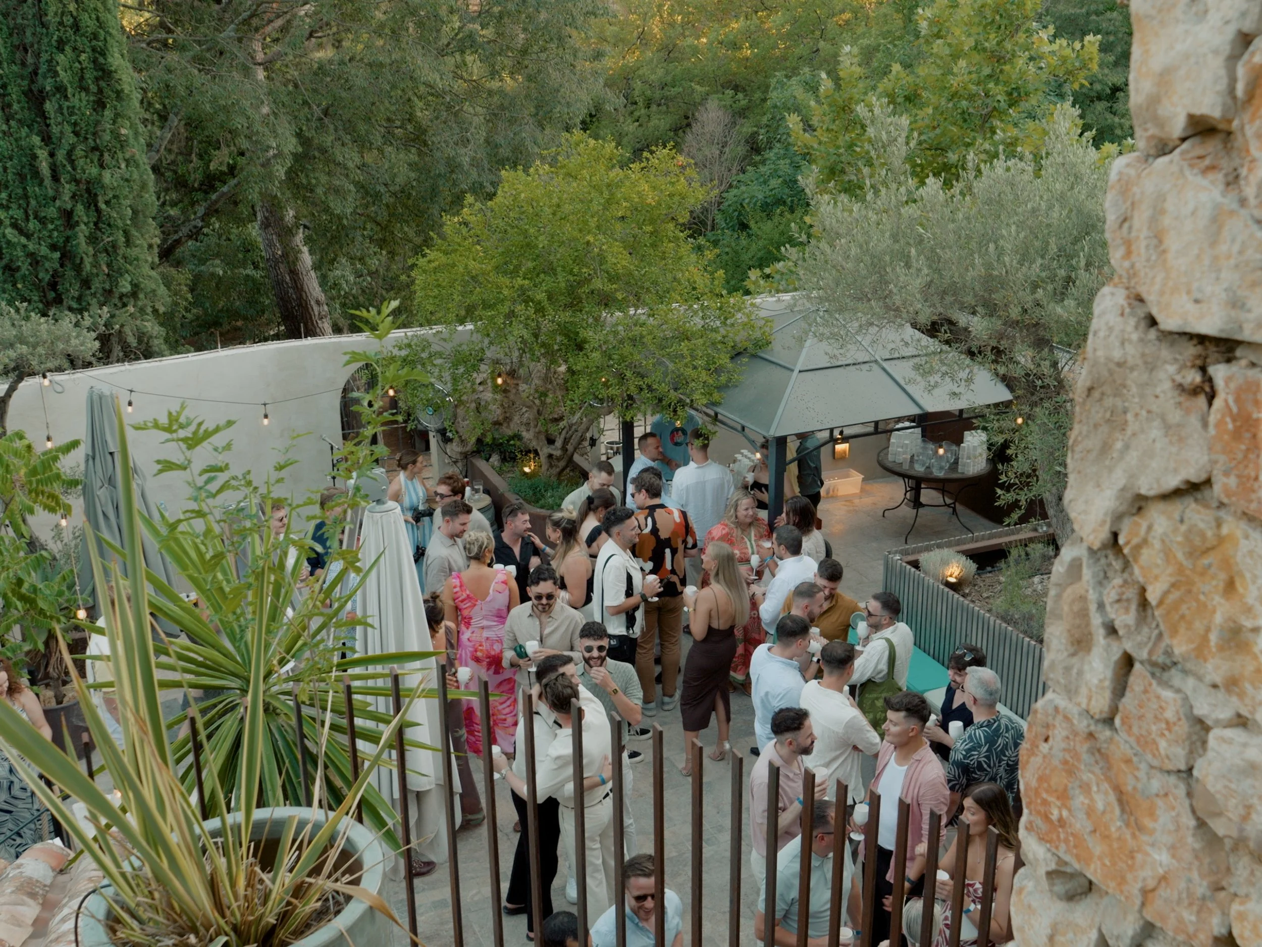 People socializing at an outdoor welcome drinks gathering before a wedding with trees and string lights in the background.
