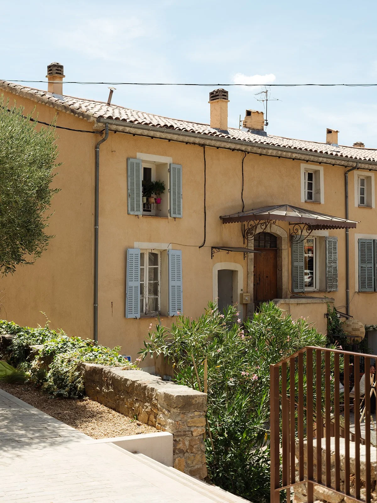 Yellow Mediterranean-style French house with blue shutters, iron awning, and a garden with green plants in Cotignac, France