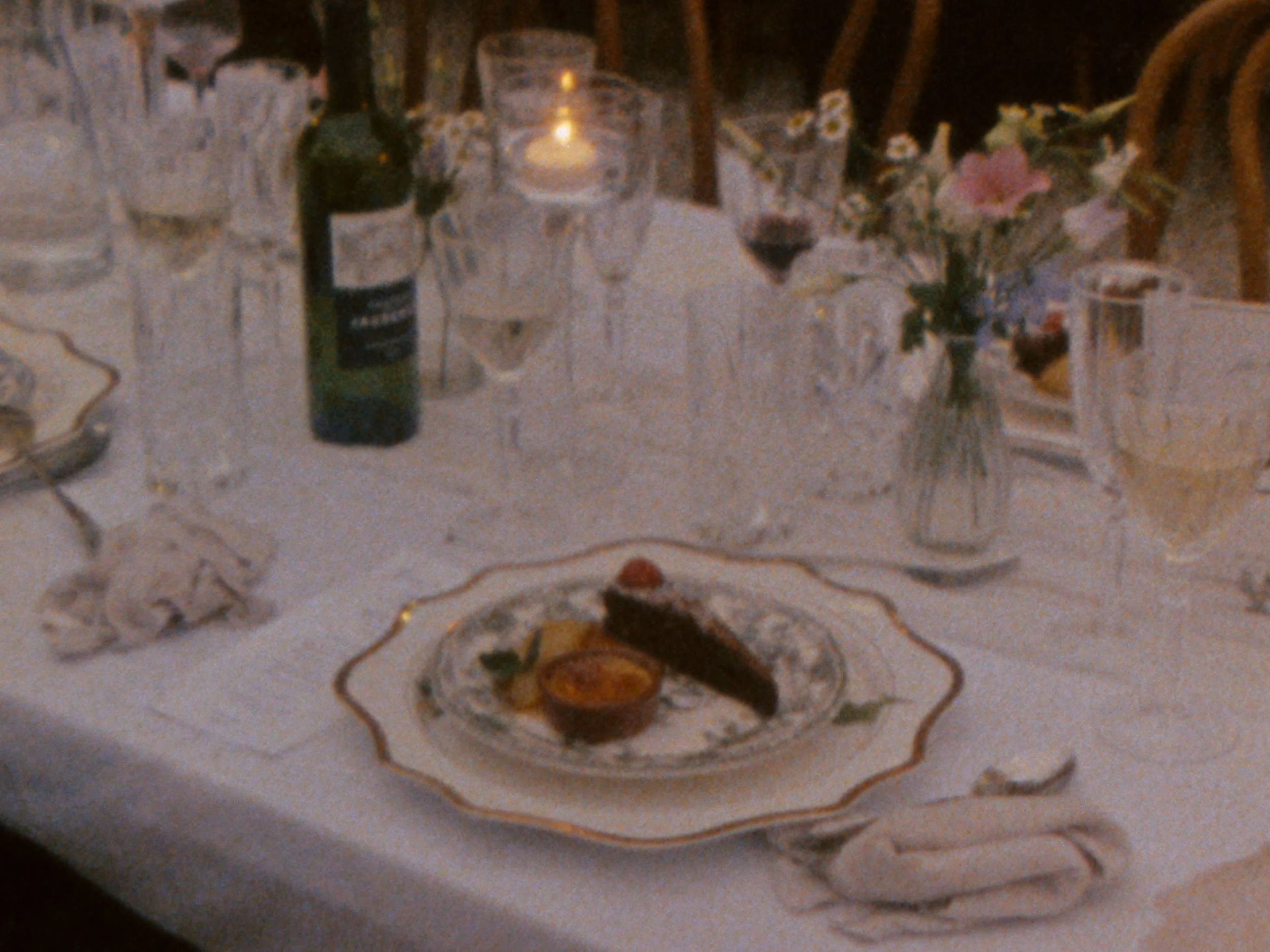 A table set for a meal at a weding, with a bottle of alcohol, a candle, a flower vase with pink flowers, wine glasses, and decorative elements on a patterned tablecloth.