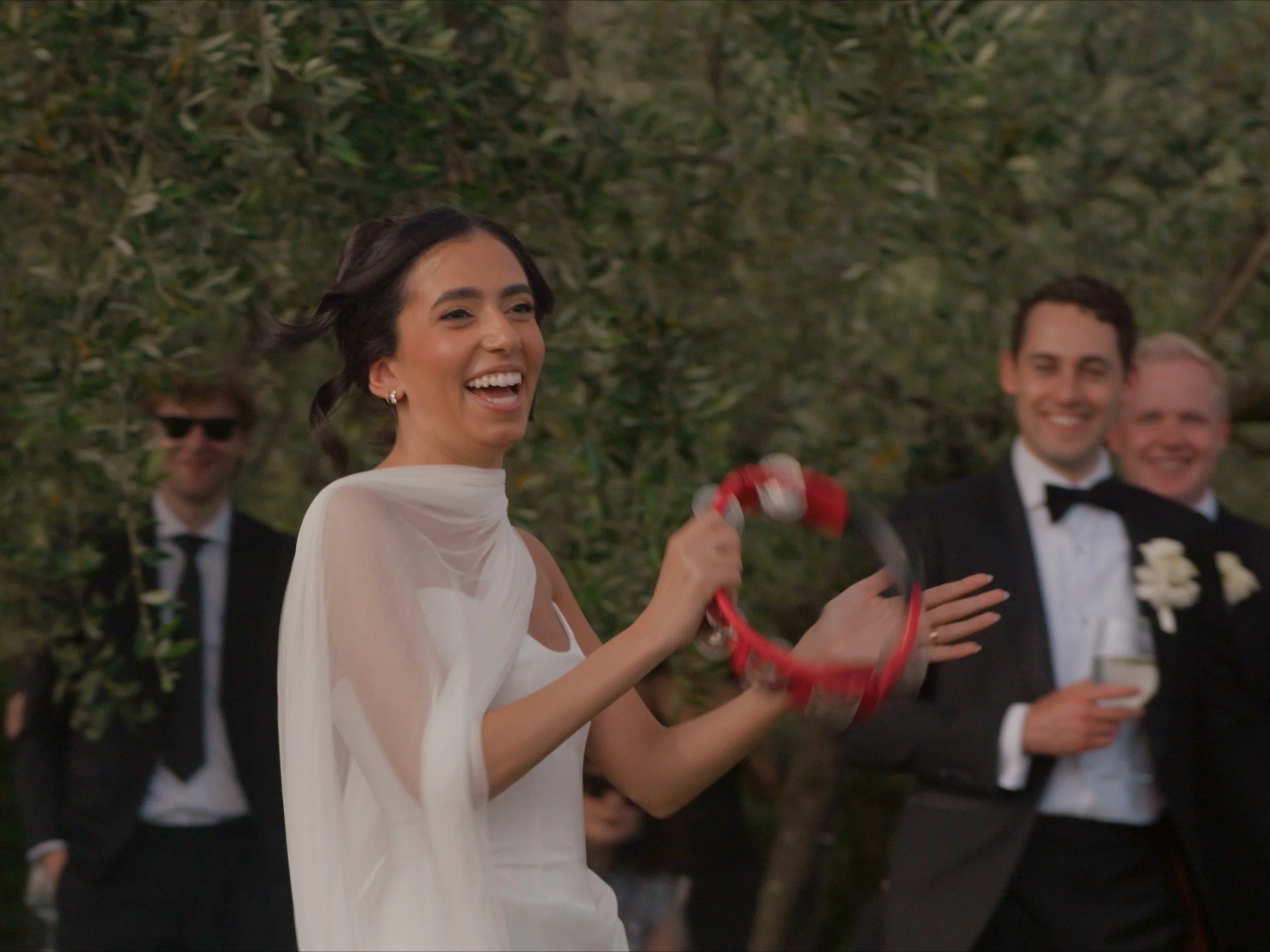 A woman in a white dress is smiling and holding red and white ribbons at an outdoor event, with three men in tuxedos and glasses in the background.