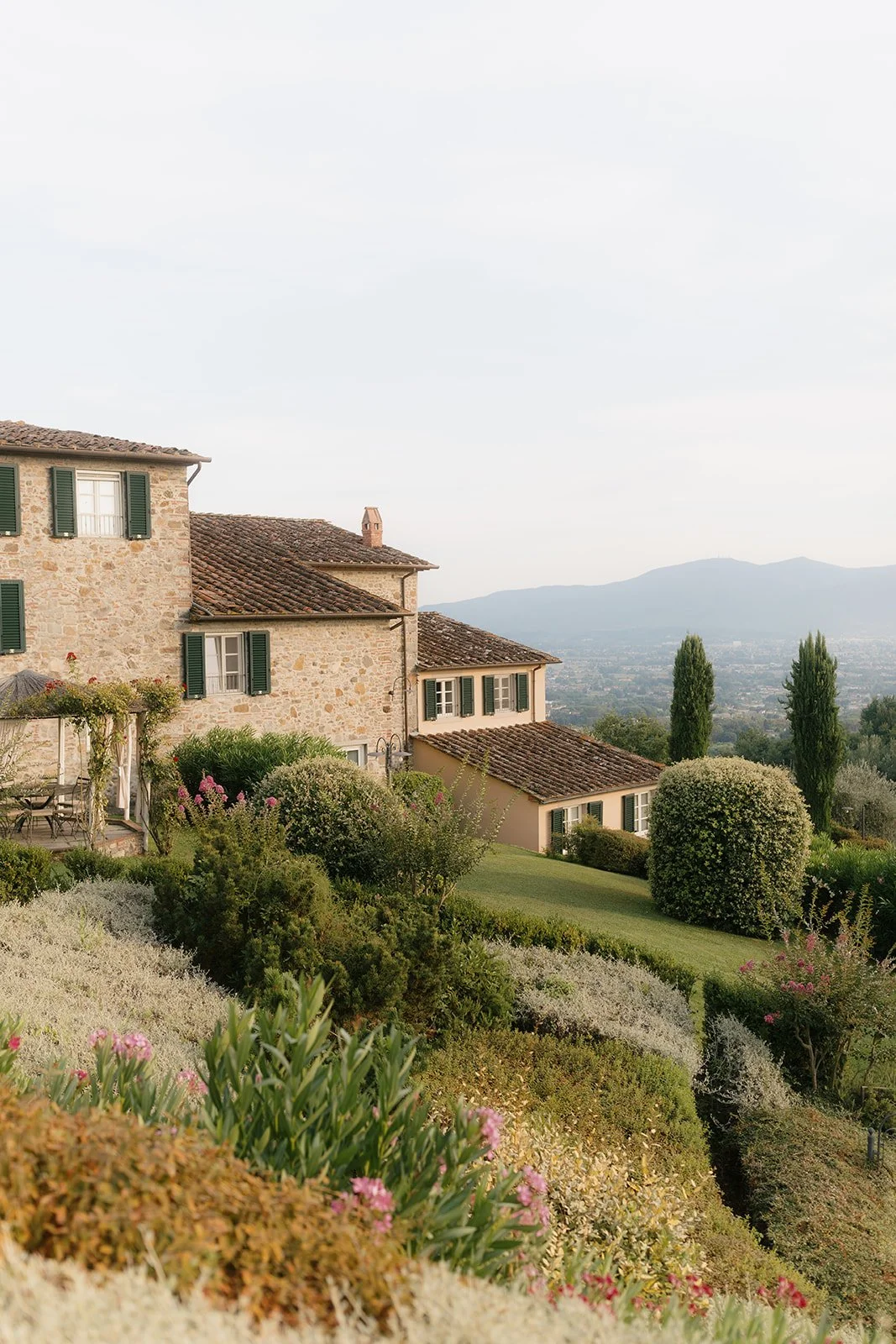 A scenic view of a rustic stone house with green shutters on a hillside, surrounded by lush garden and colorful flowering bushes, with distant mountains and a soft cloudy sky in the background.