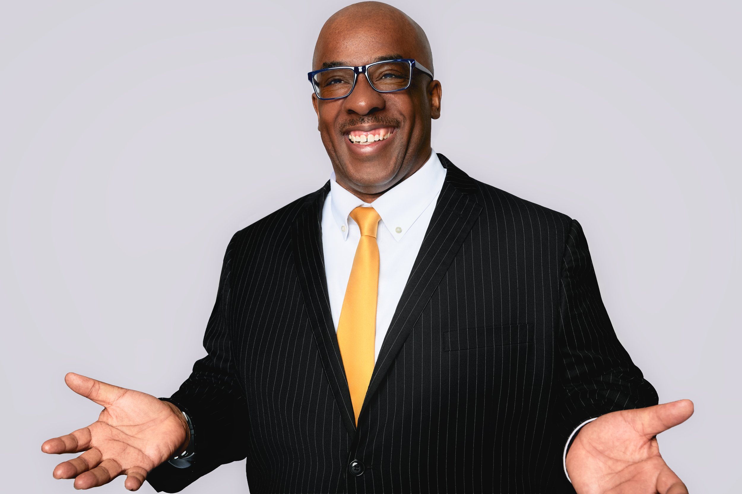 A smiling man in a black pinstripe suit with a white shirt and yellow tie is gesturing with open hands against a plain light gray background.