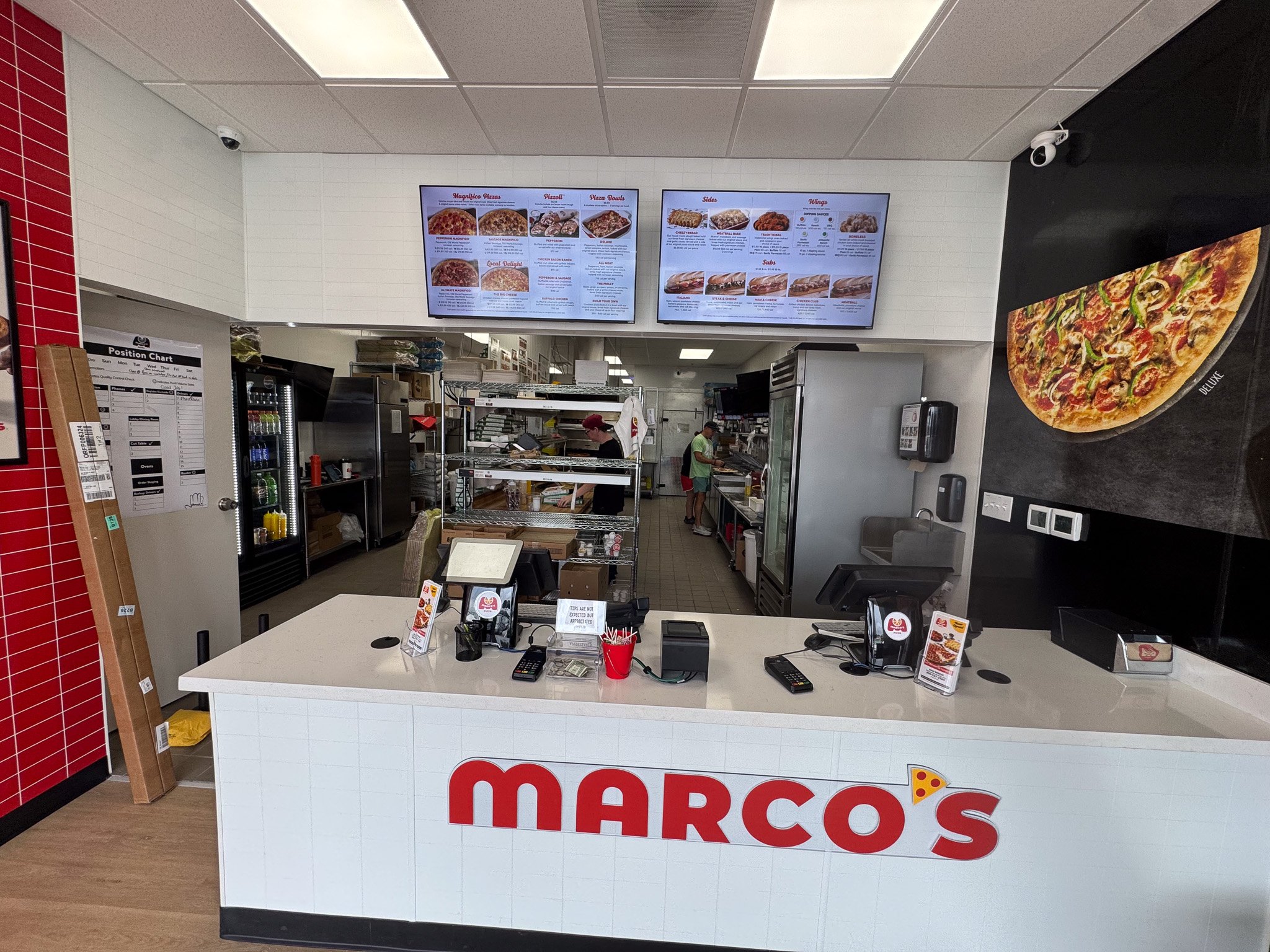 Quick service restaurant counter with digital menu screens displaying pizza options, staff preparing food in the background, and a large pizza advertisement sign on the right side.