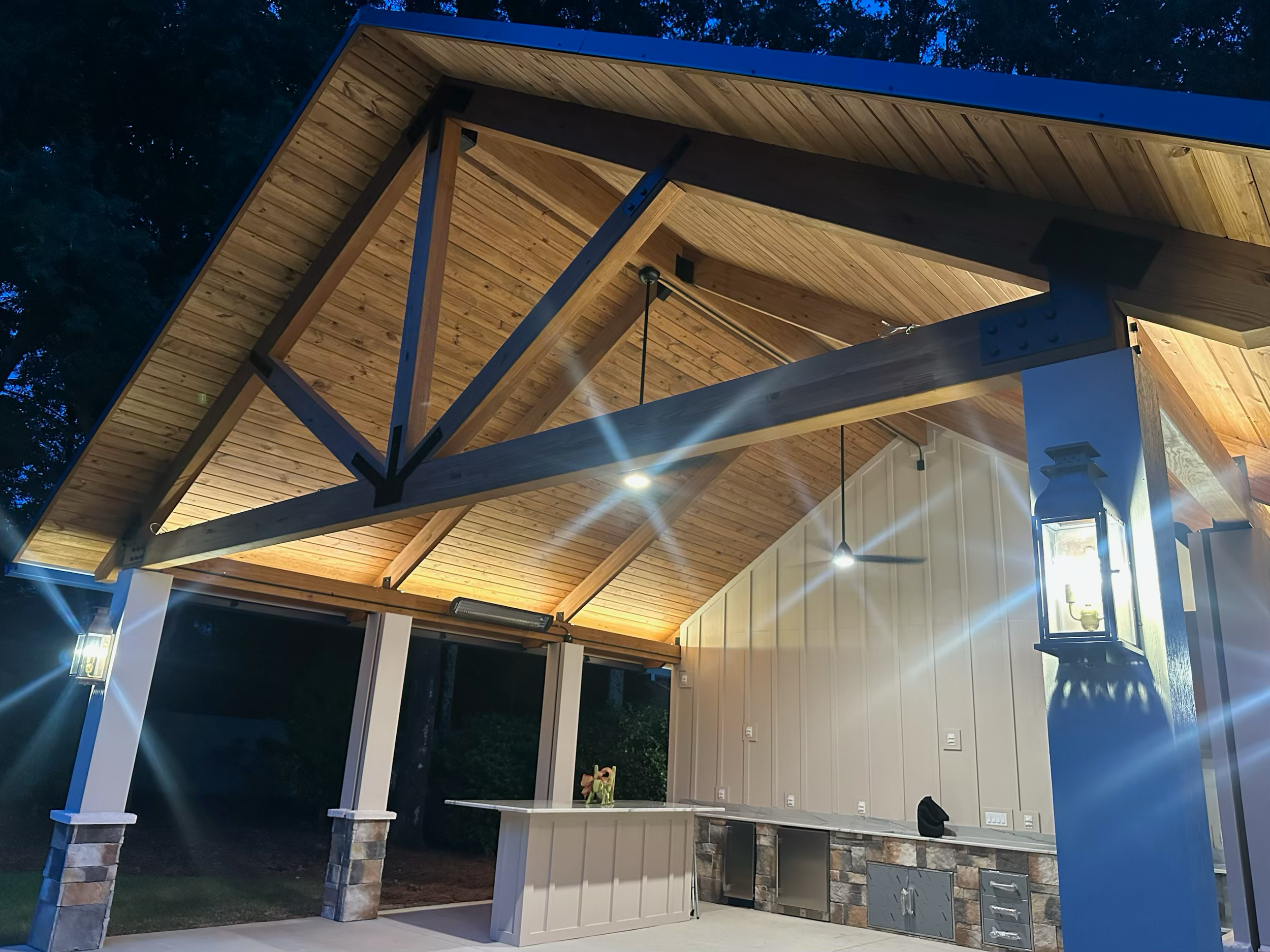 Backyard patio area with a wooden pergola roof, string lights, and a counter with stone base and cabinetry. There is a small decorative item on the counter and a lantern hanging on the side. The area is illuminated at night with trees visible in the background.