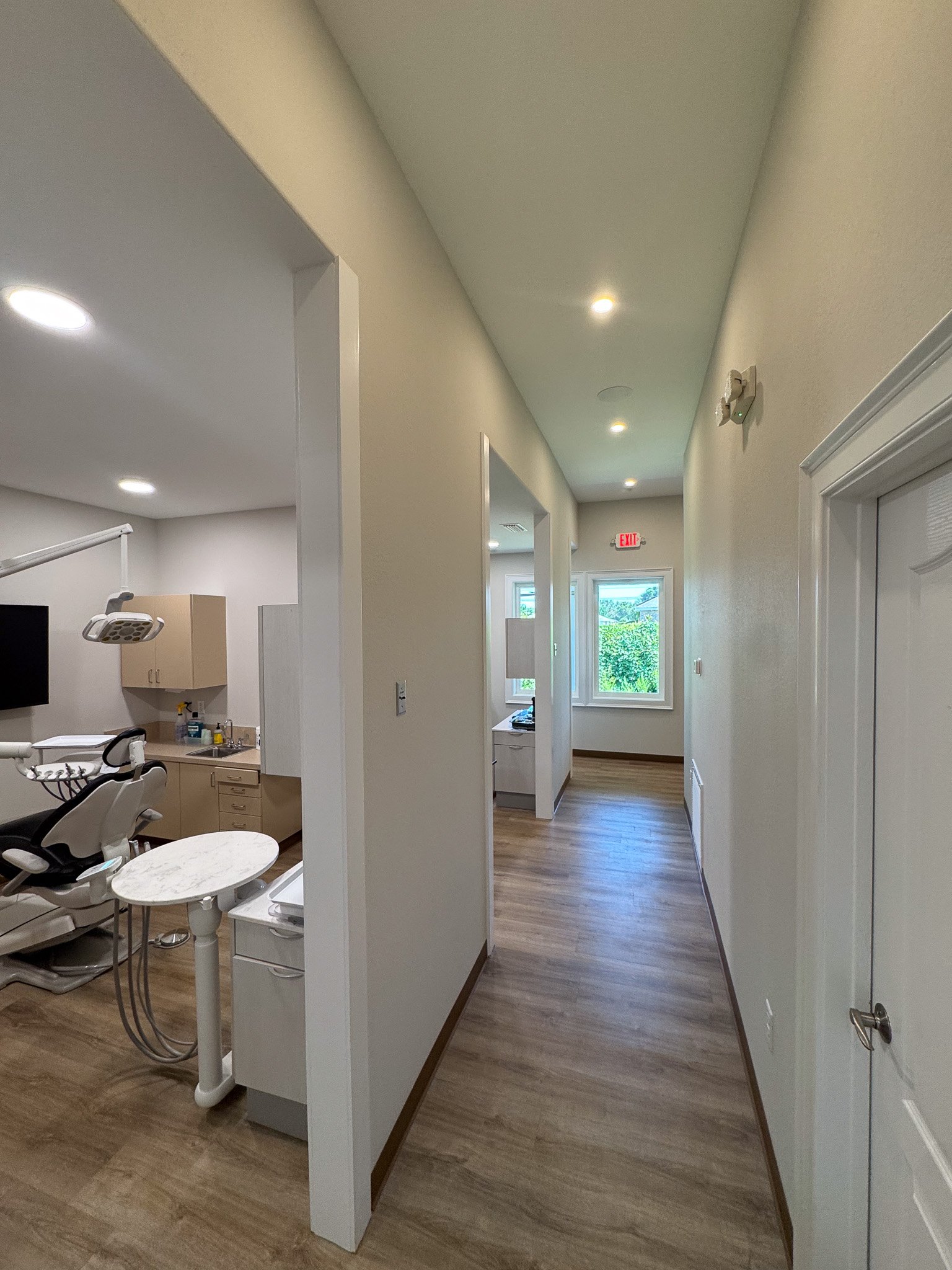 Corridor of a building with wooden flooring, white walls, and ceiling. There are multiple recessed lights and large windows at the end showing trees outside. On the left, a room with a dental chair, equipment, and a monitor is visible.