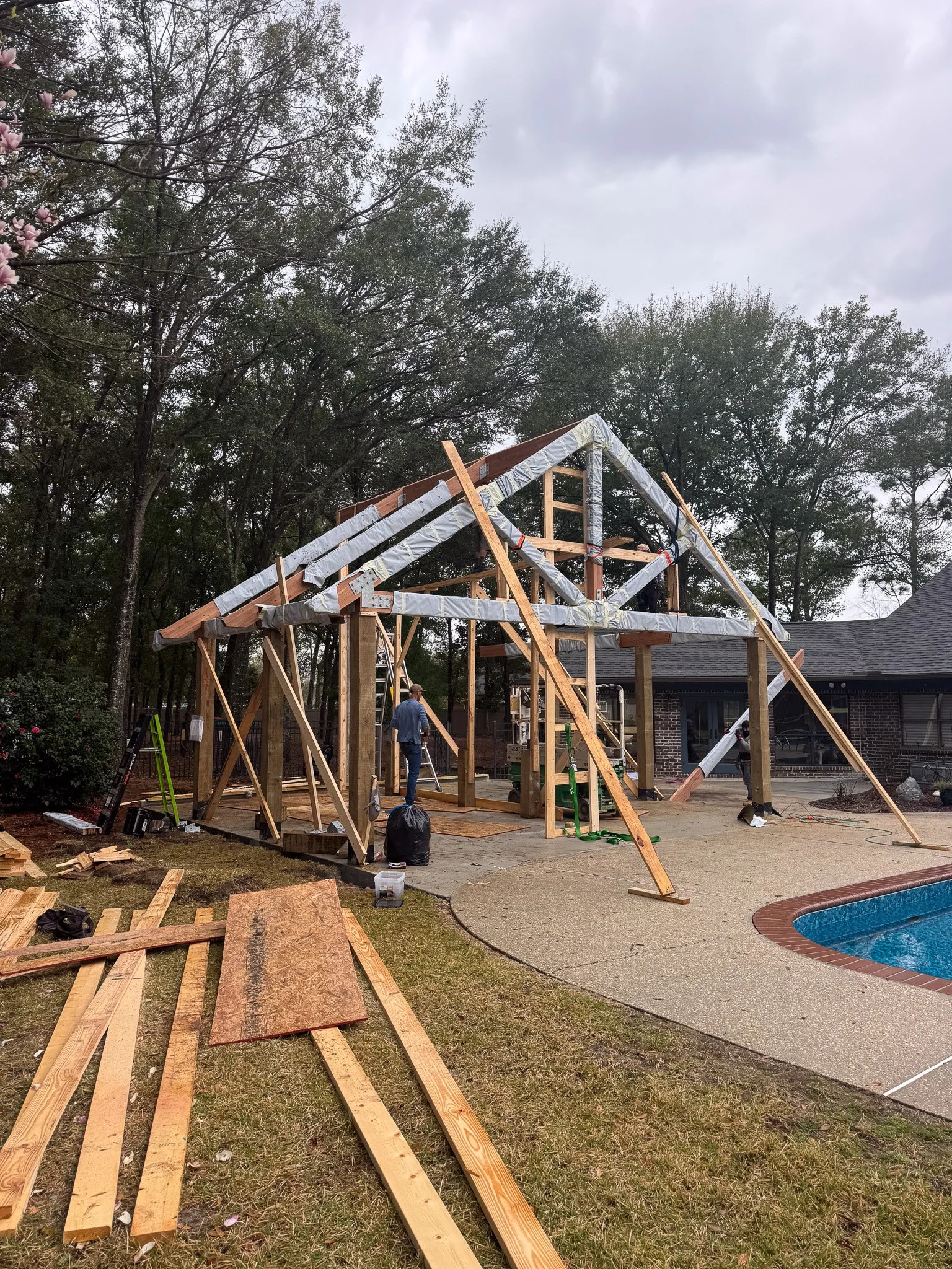 Construction of a wooden house frame in a backyard with trees, cloudy sky, and a pool nearby. Workers are assembling the structure with tools and materials scattered around.