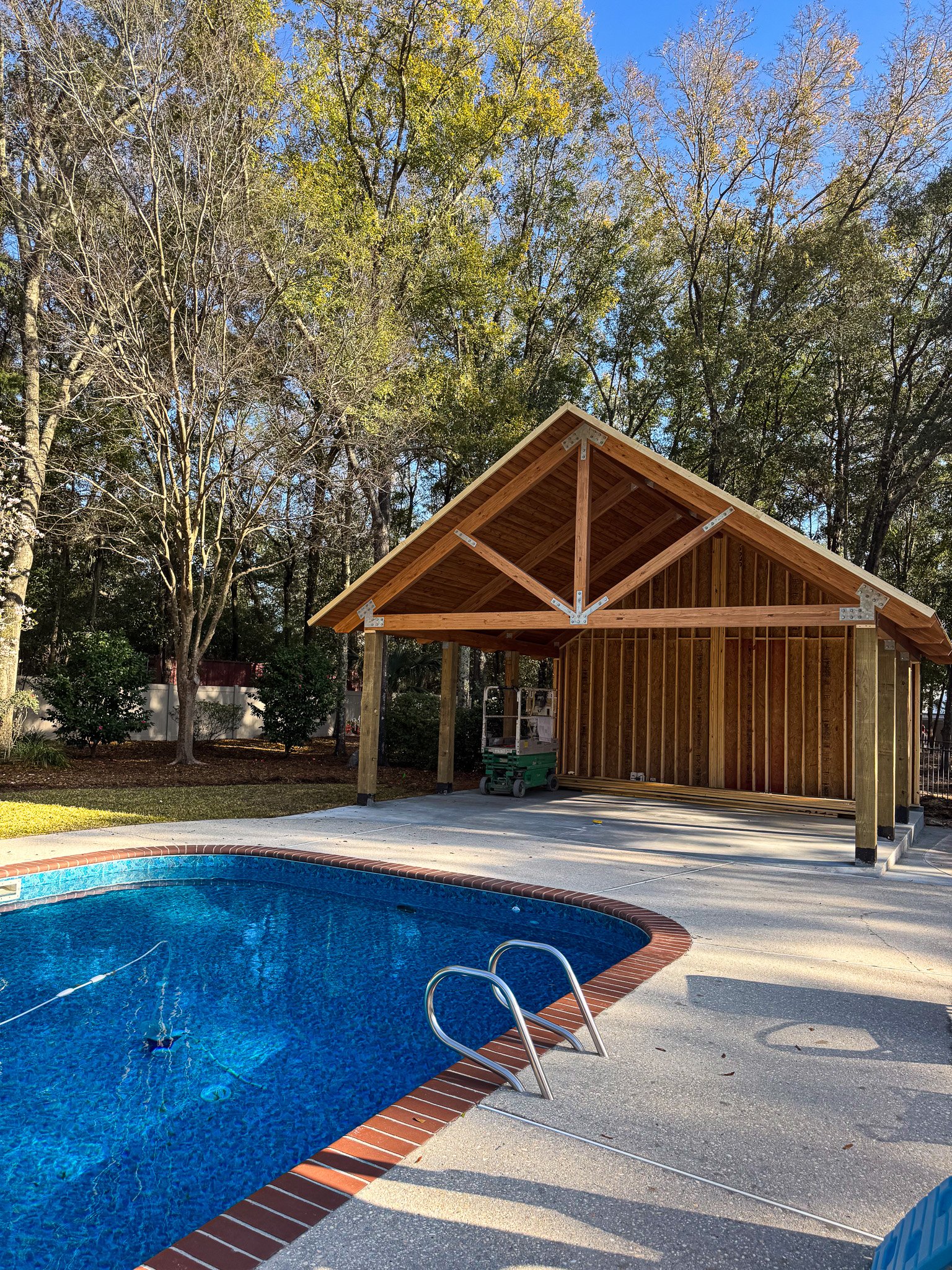 A backyard scene featuring a kidney-shaped swimming pool with blue water, a red brick coping, and a concrete pool deck. In the background, there is a wooden structure under construction with exposed framing and a gable roof, surrounded by trees with autumn foliage and a clear blue sky.