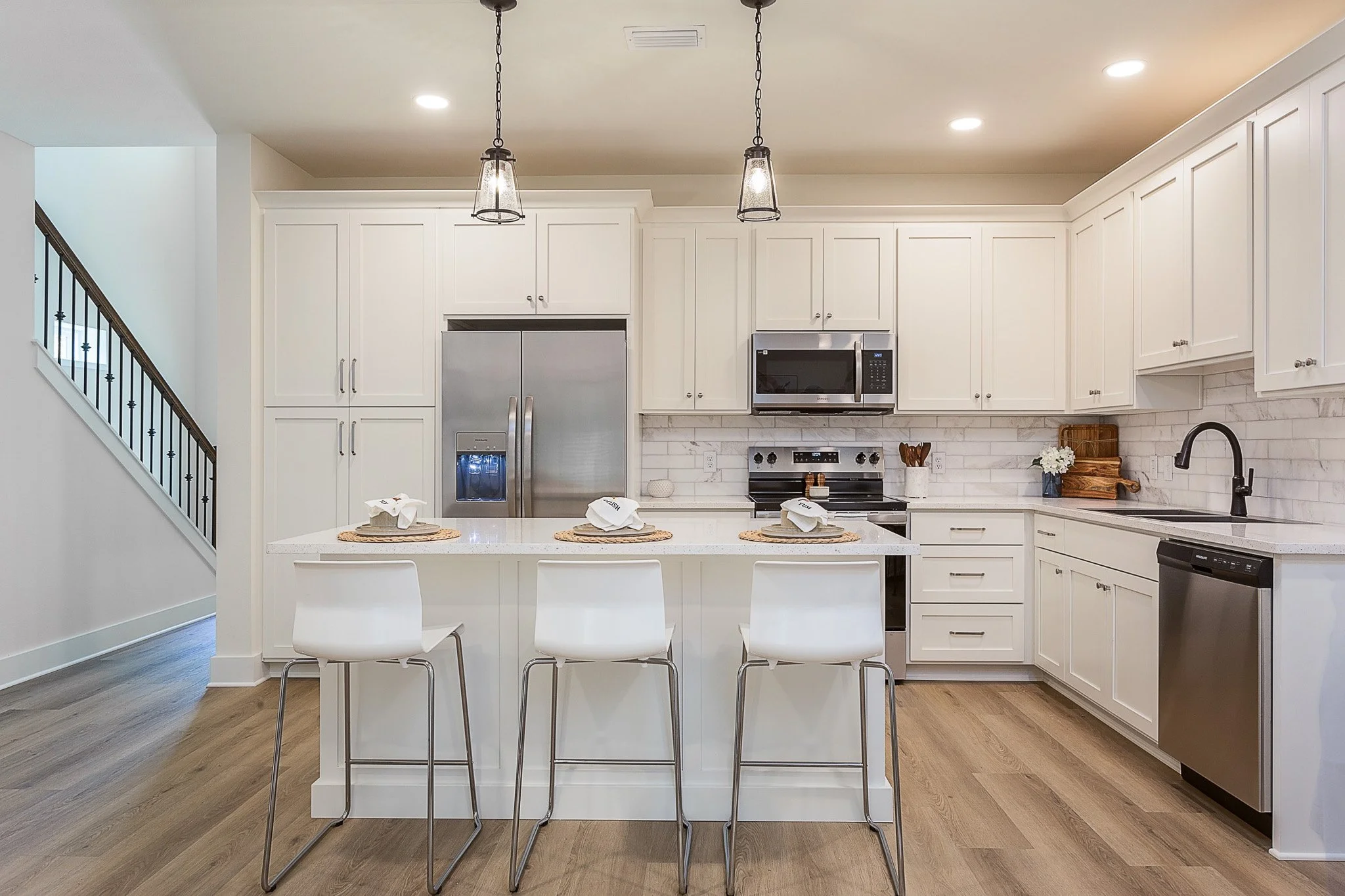 Modern kitchen with white cabinets, stainless steel appliances, and a white island with four white chairs. There are pendant lights hanging above the island, and a staircase to the left.
