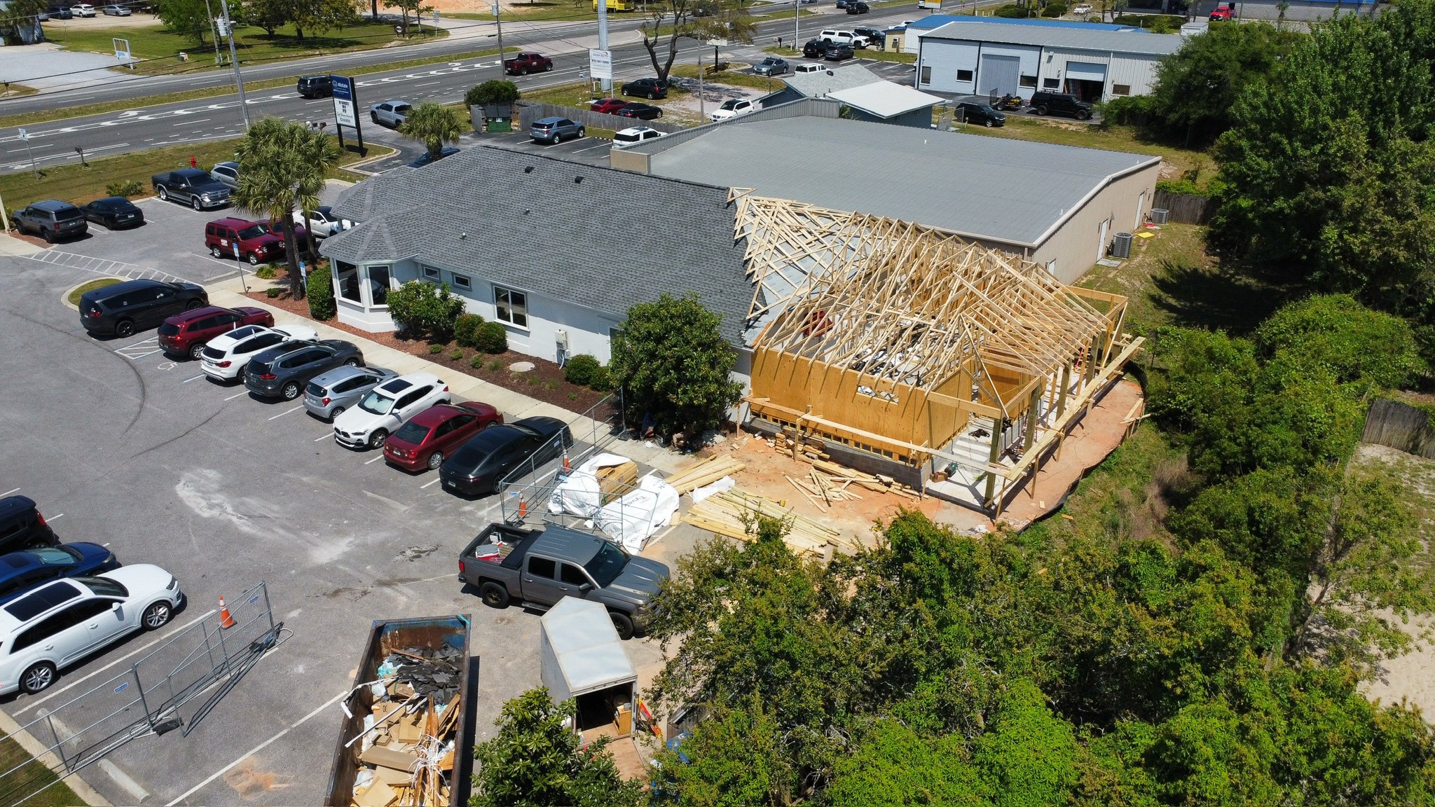 Aerial view of a building under construction with a wooden framework. Attached to a parking lot with numerous parked cars, near a busy road with cars and traffic signals. Green trees surround the area.