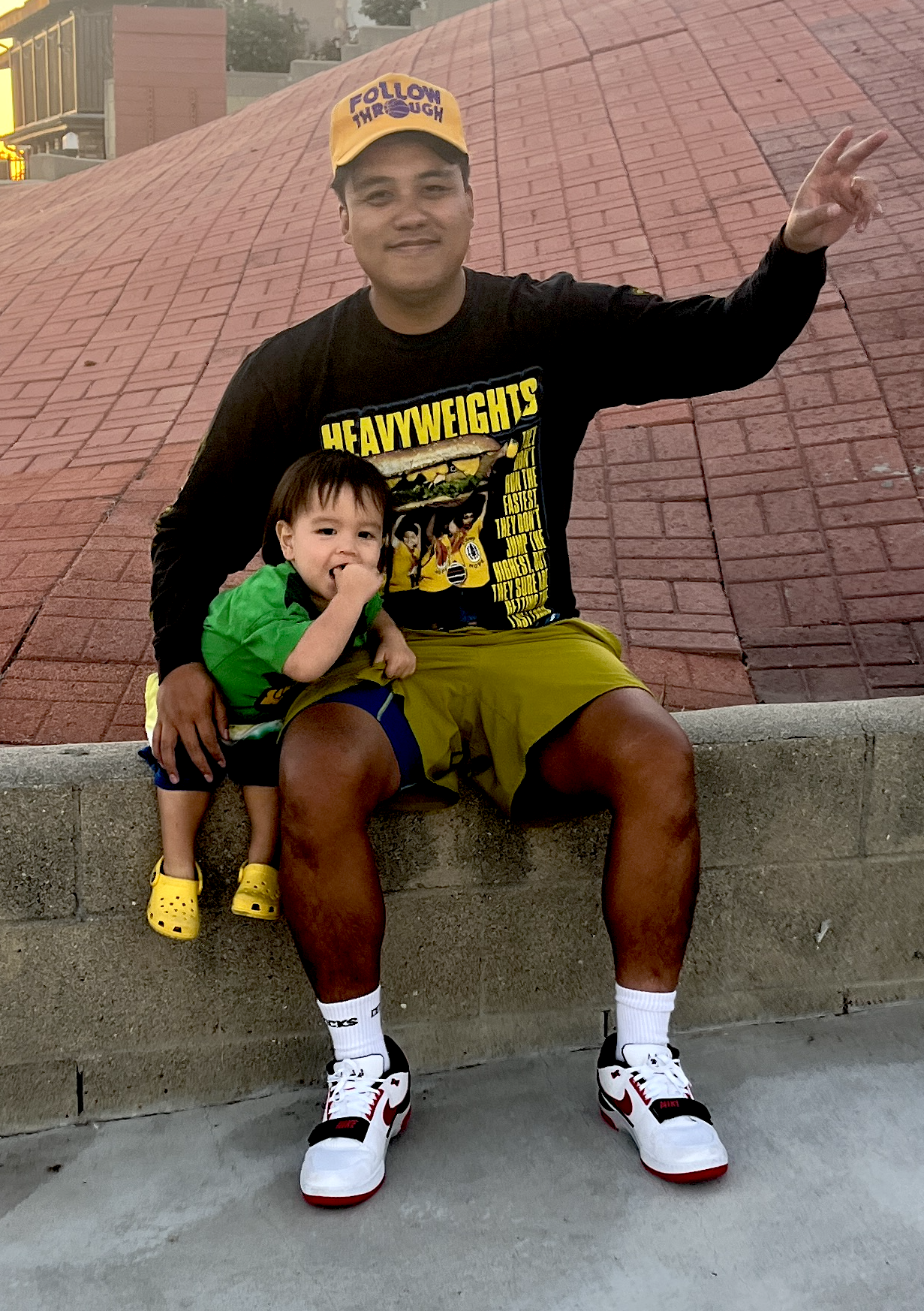 A man and a young boy sitting on a concrete ledge outdoors. The man is wearing a yellow cap with purple text, a black long-sleeve shirt with a comic-style graphic and text, yellow shorts, white socks, and white and red sneakers. The young boy is wearing a green shirt, navy shorts with yellow accents, yellow Crocs, and is sitting on the man’s lap, sucking his thumb. The background features a red brick wall and some urban buildings.